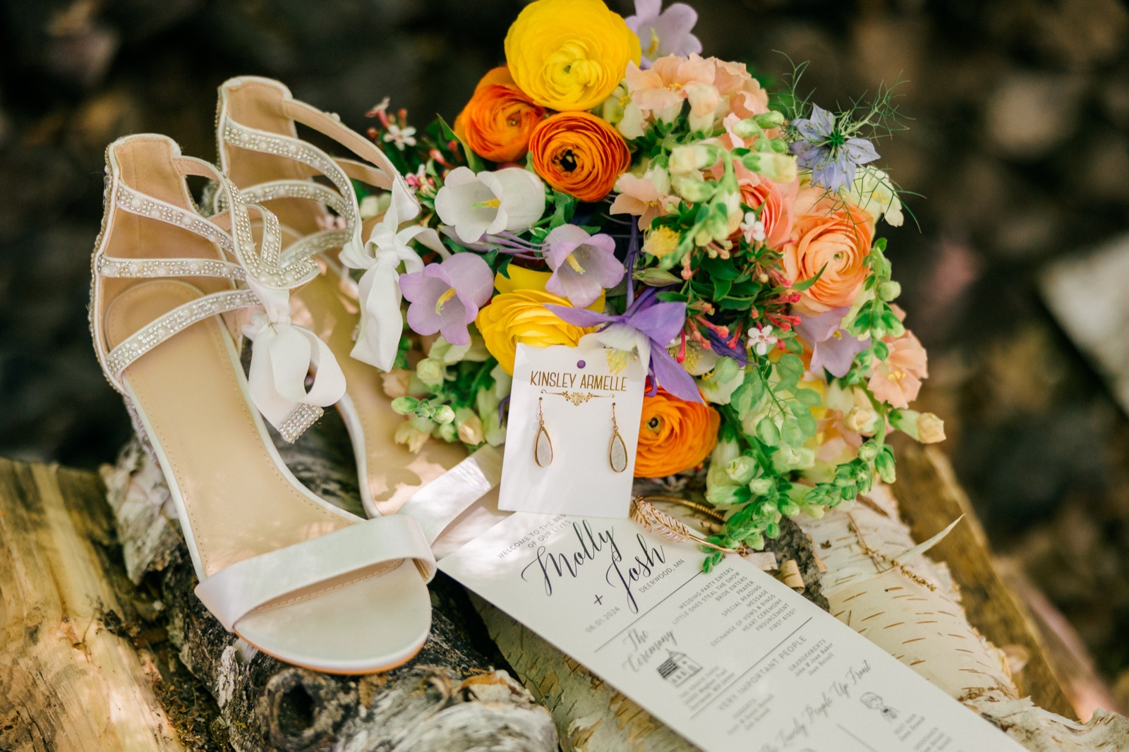 A bridal lay-flat on birch logs featuring strappy ivory heels, a vibrant bouquet of ranunculus and snapdragons in citrus and pastel tones, teardrop earrings, and a ceremony program — Tim Larsen Photography, Brainerd Lakes MN