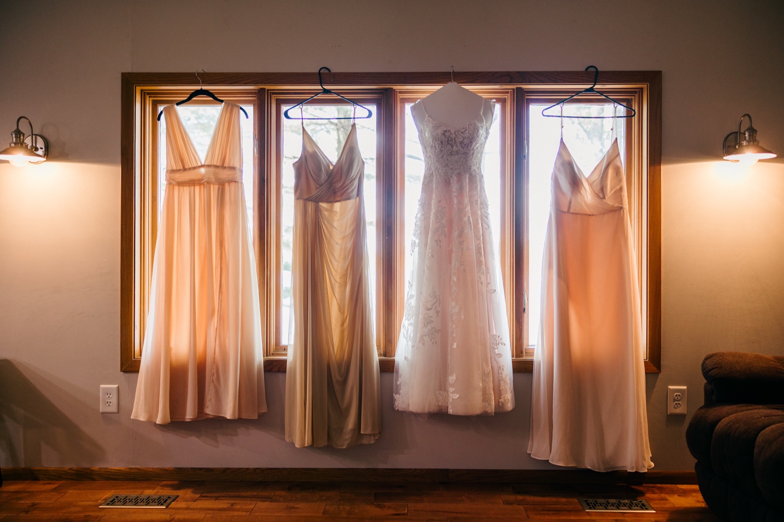 The bride's gown and three bridesmaid dresses hang backlit in a row of cabin windows flanked by wall sconces — Tim Larsen Photography, Brainerd Lakes MN