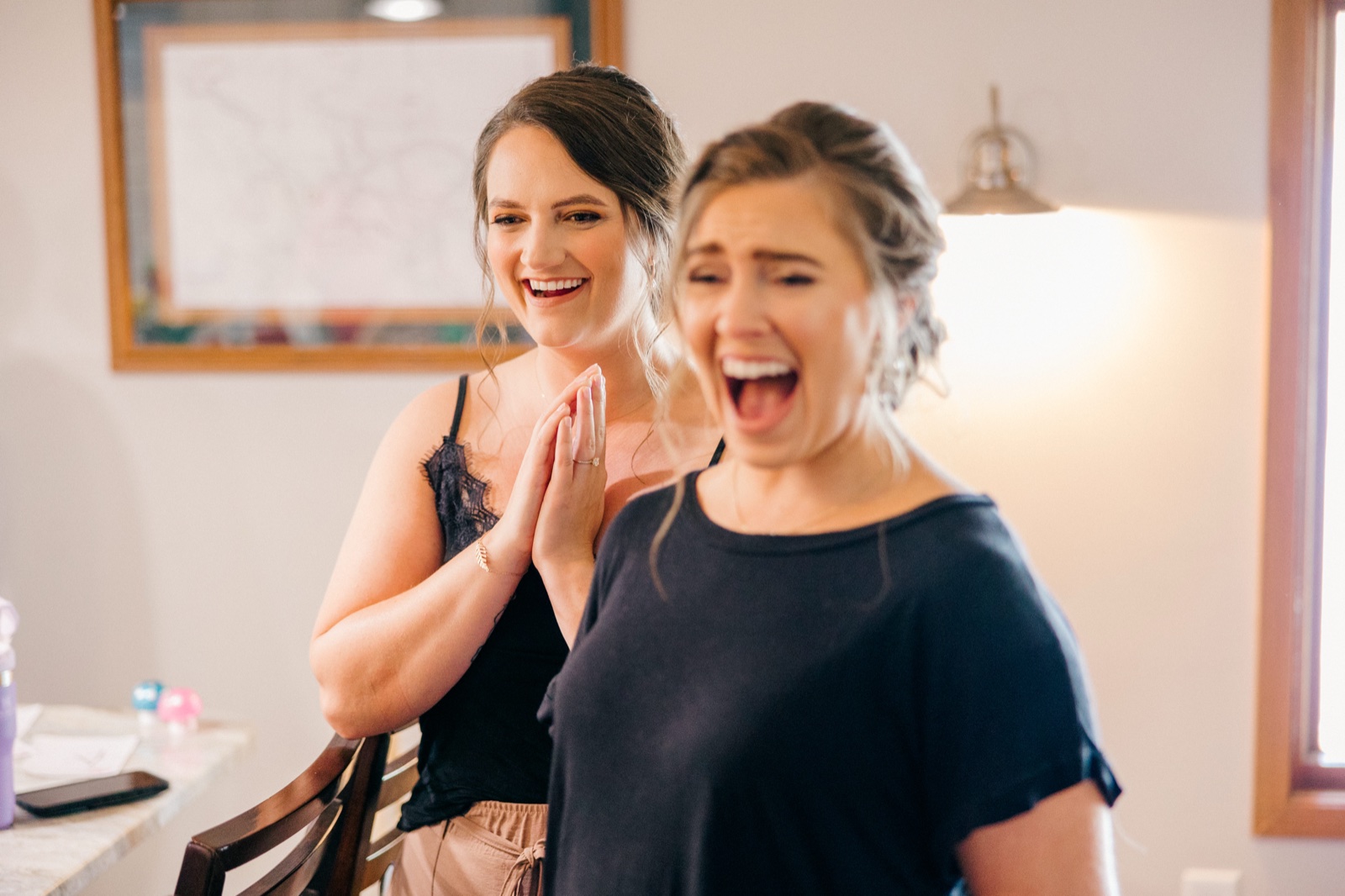 A bridesmaid presses her hands together and laughs while another woman in the foreground reacts with an open-mouthed laugh during getting-ready inside the cabin — Tim Larsen Photography, Brainerd Lakes MN