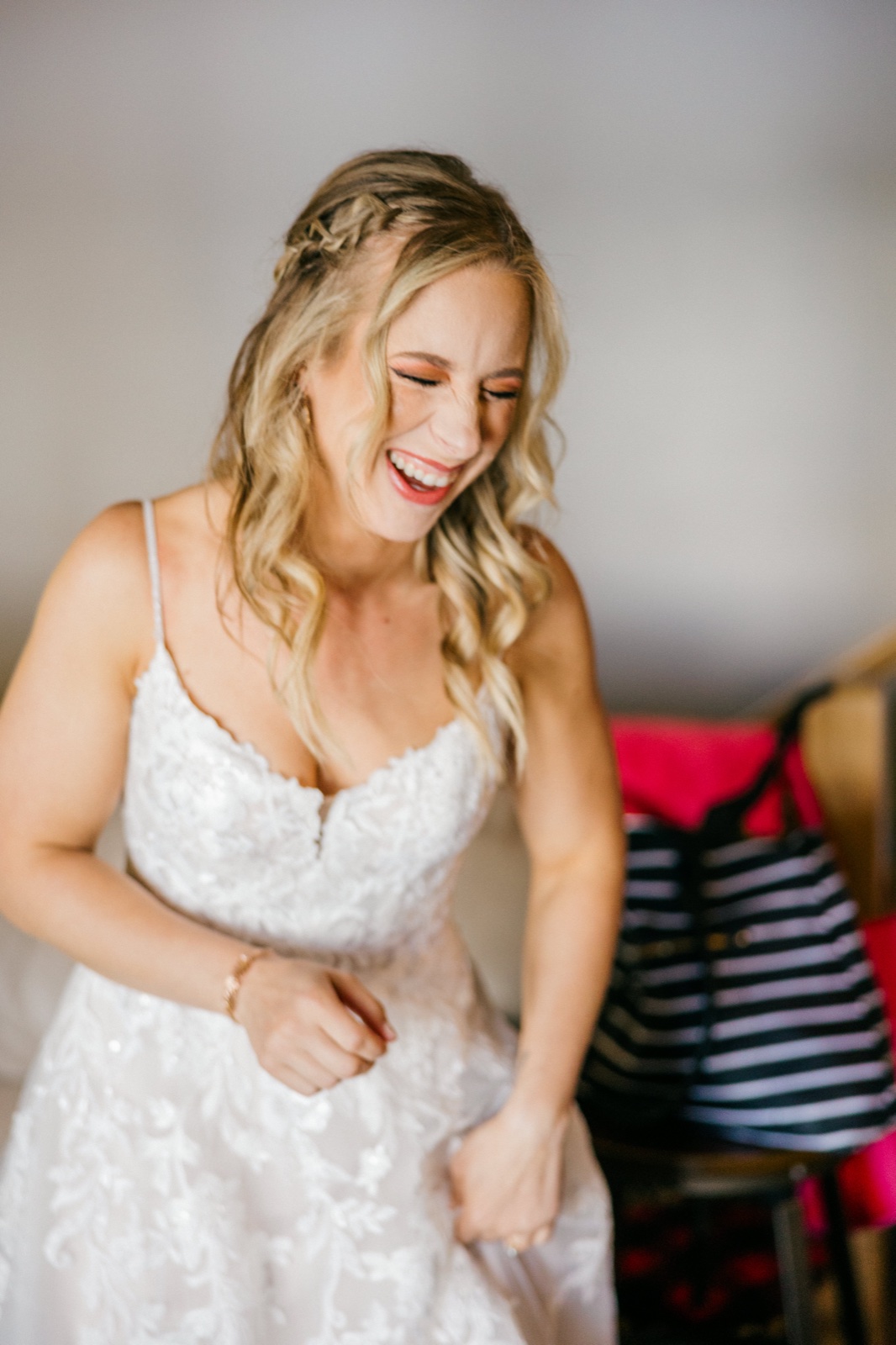 The bride throws her head back laughing while holding the skirt of her lace wedding gown in a softly lit getting-ready room — Tim Larsen Photography, Brainerd Lakes MN