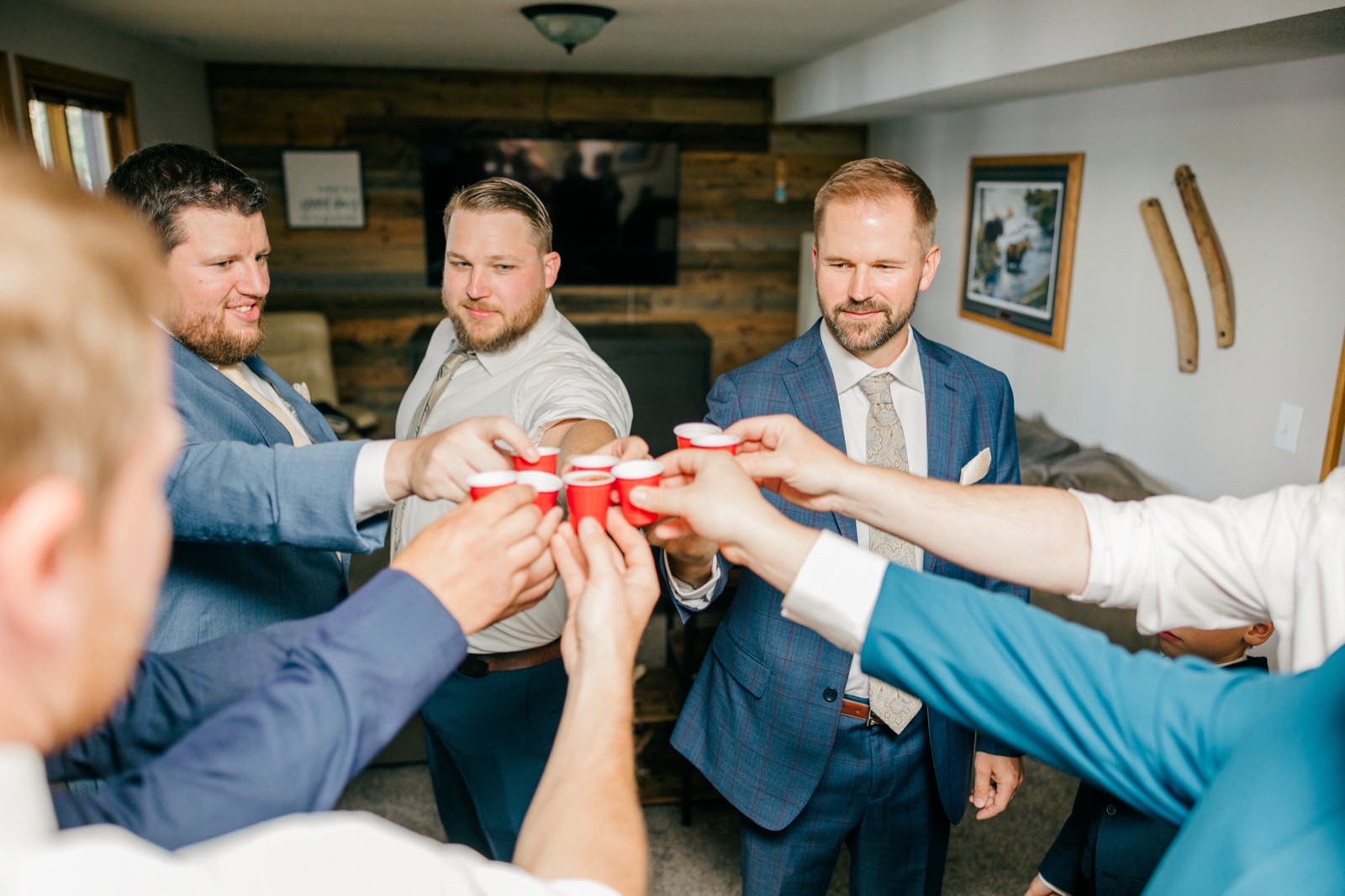 The groom and his groomsmen extend their arms inward to clink small red plastic shot cups together in a wood-paneled cabin lounge — Tim Larsen Photography, Brainerd Lakes MN