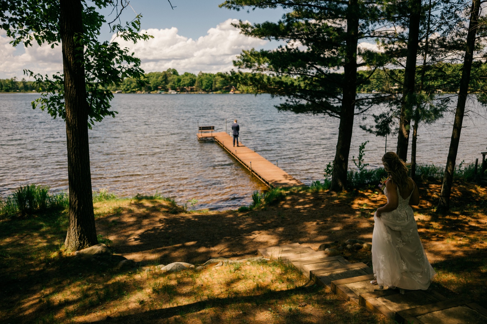 The bride steps down a wooden path toward the lake while the groom waits with his back turned at the end of a long dock for the first look — Tim Larsen Photography, Brainerd Lakes MN