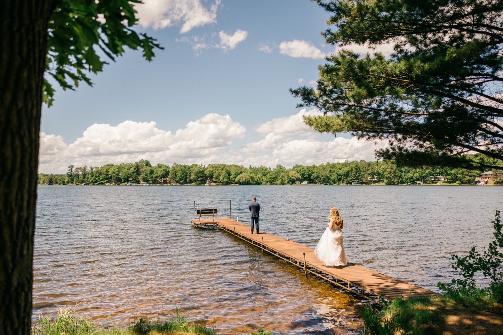 Framed by overhanging tree branches, the bride walks down the long wooden dock toward the groom waiting at the end above the calm Brainerd Lakes-area lake — Tim Larsen Photography, Brainerd Lakes MN