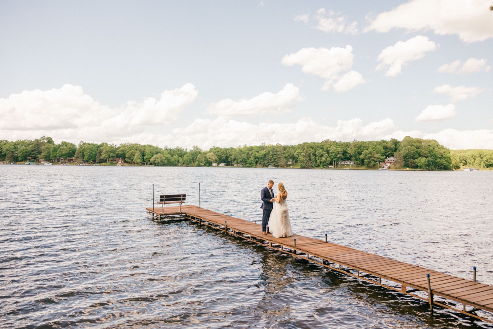 The couple stands together at the end of a long wooden dock extending into a Brainerd Lakes-area lake, reading vows beneath a wide cloud-filled sky — Tim Larsen Photography, Brainerd Lakes MN