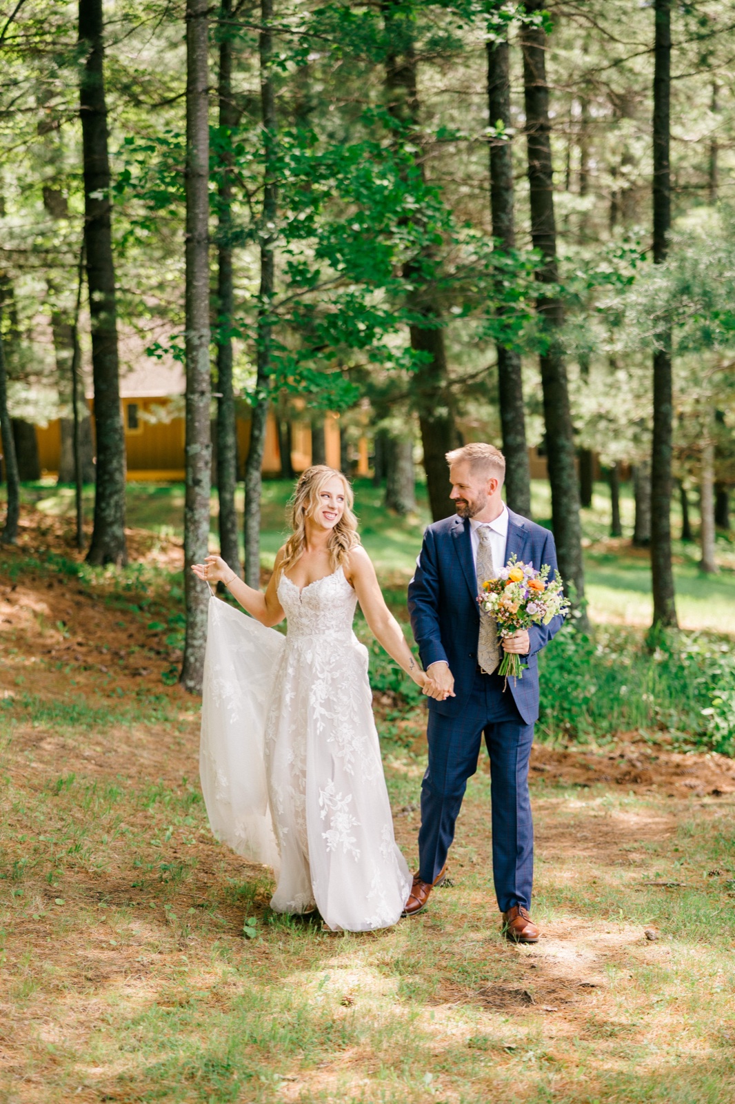 The bride lifts her gown and laughs at the groom as they walk hand-in-hand through a sunlit pine forest, a colorful bouquet at his side — Tim Larsen Photography, Brainerd Lakes MN