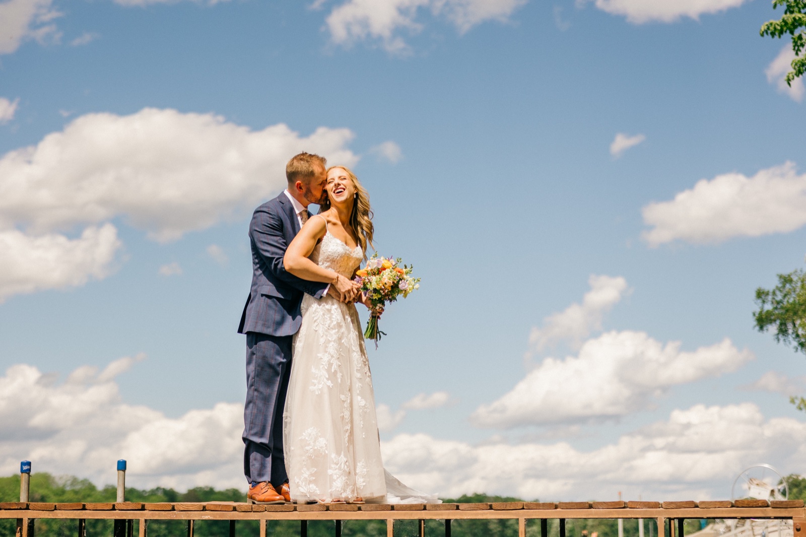 The groom kisses the bride's temple as she laughs and holds a colorful bouquet, the two standing on a wooden dock beneath a vast blue sky filled with summer clouds — Tim Larsen Photography, Brainerd Lakes MN