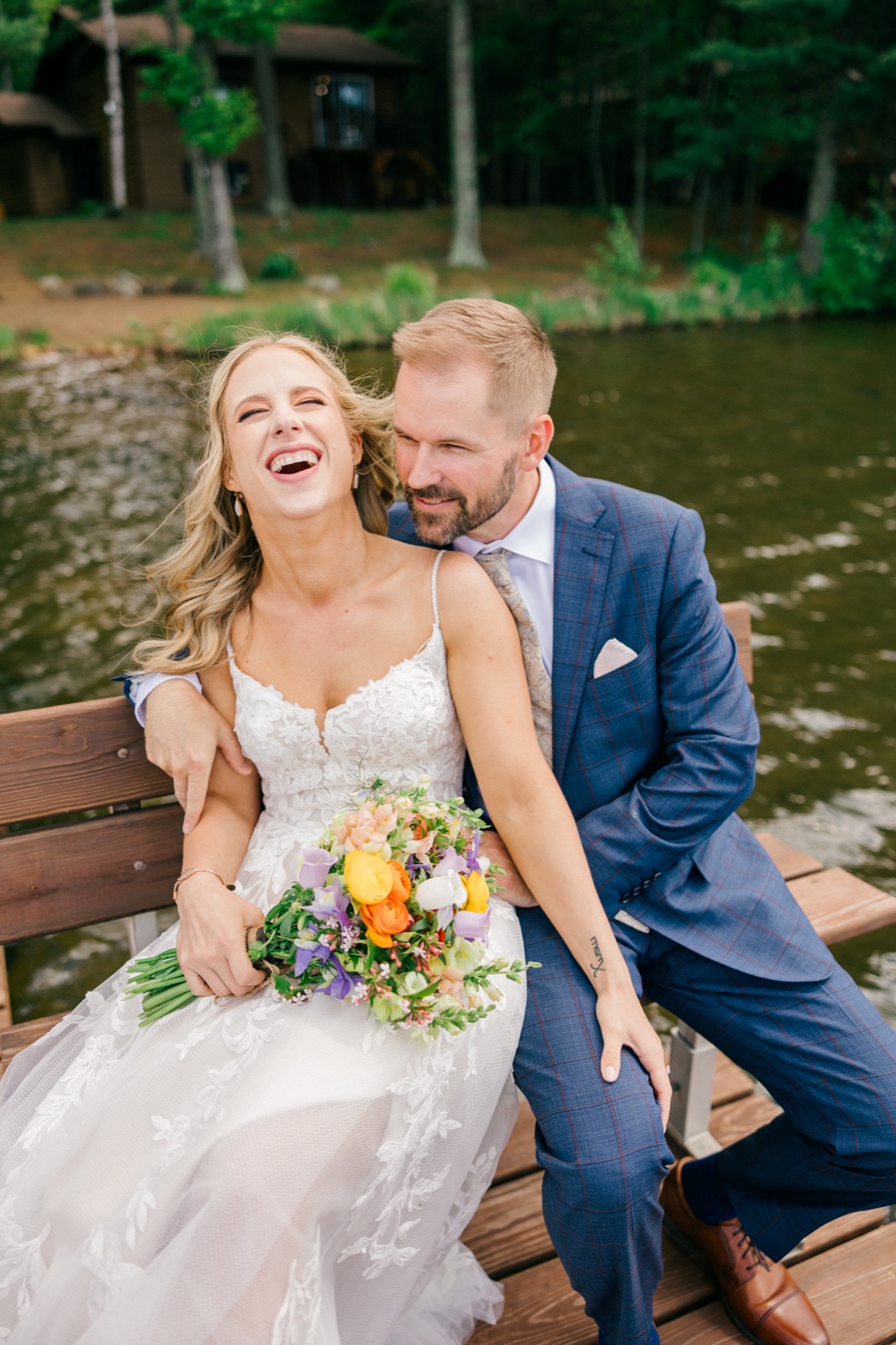 The bride throws her head back laughing while the groom leans in close, the two seated on a wooden bench at the edge of the lake with a cabin visible behind them — Tim Larsen Photography, Brainerd Lakes MN