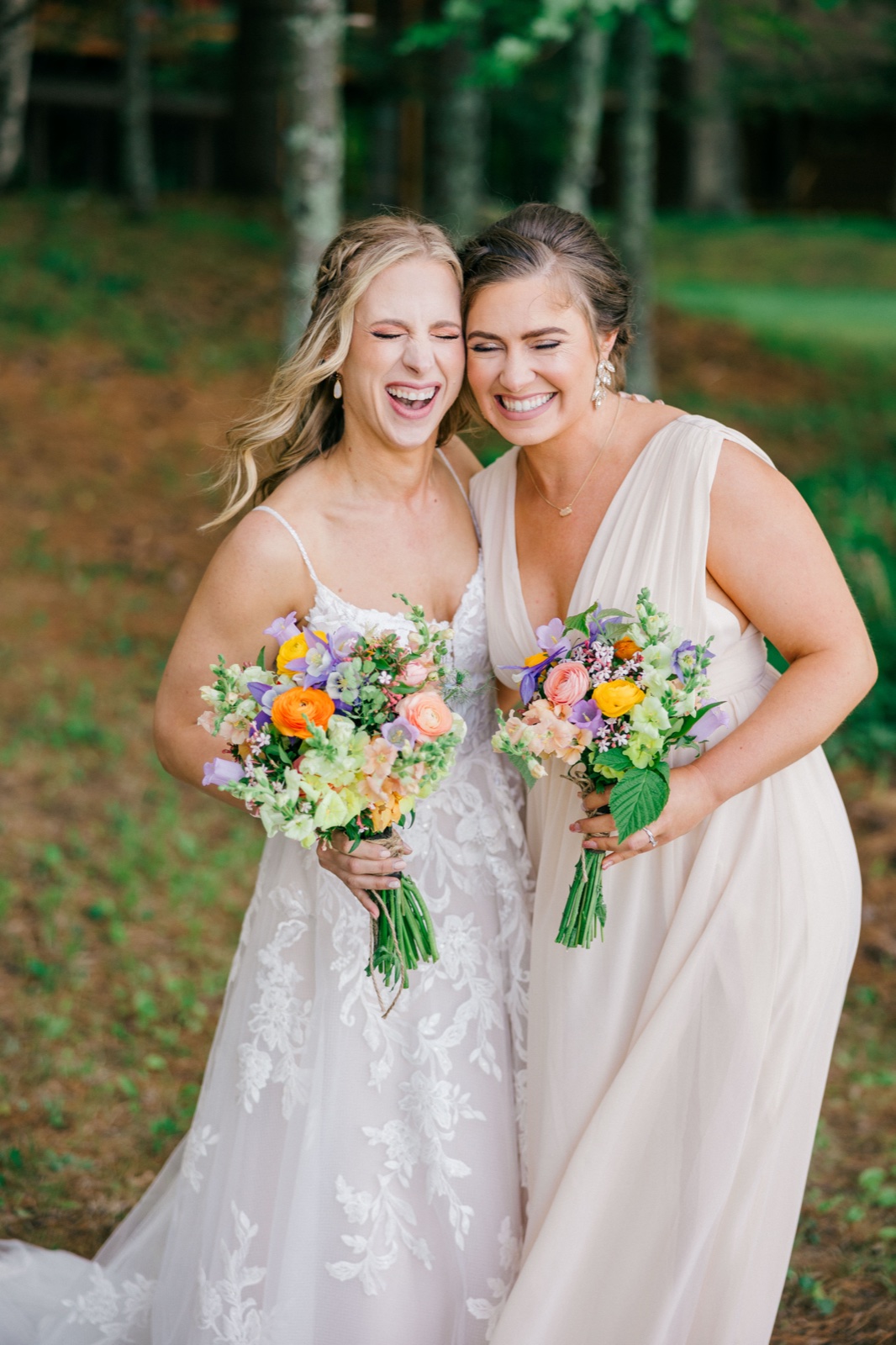 The bride and a bridesmaid lean cheek-to-cheek laughing wide-mouthed while holding colorful wildflower bouquets in a wooded clearing — Tim Larsen Photography, Brainerd Lakes MN