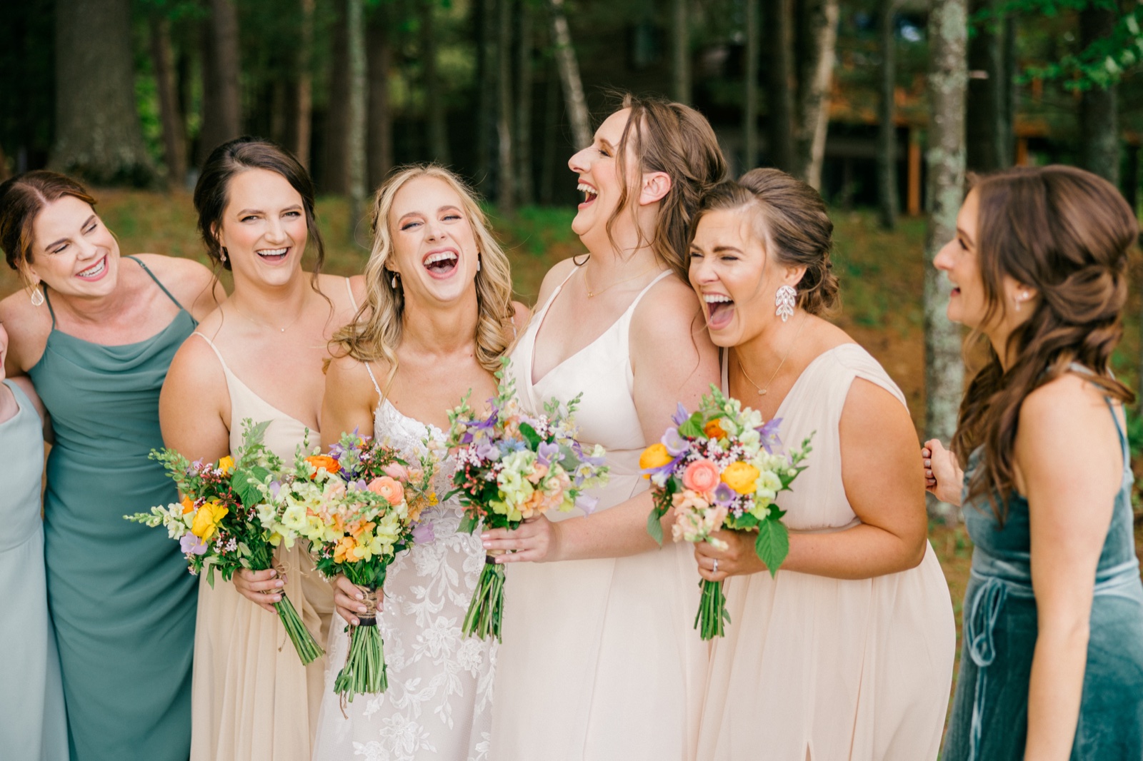 The bride and her five bridesmaids laugh together holding colorful spring bouquets on a wooded lawn outside the cabin — Tim Larsen Photography, Brainerd Lakes MN