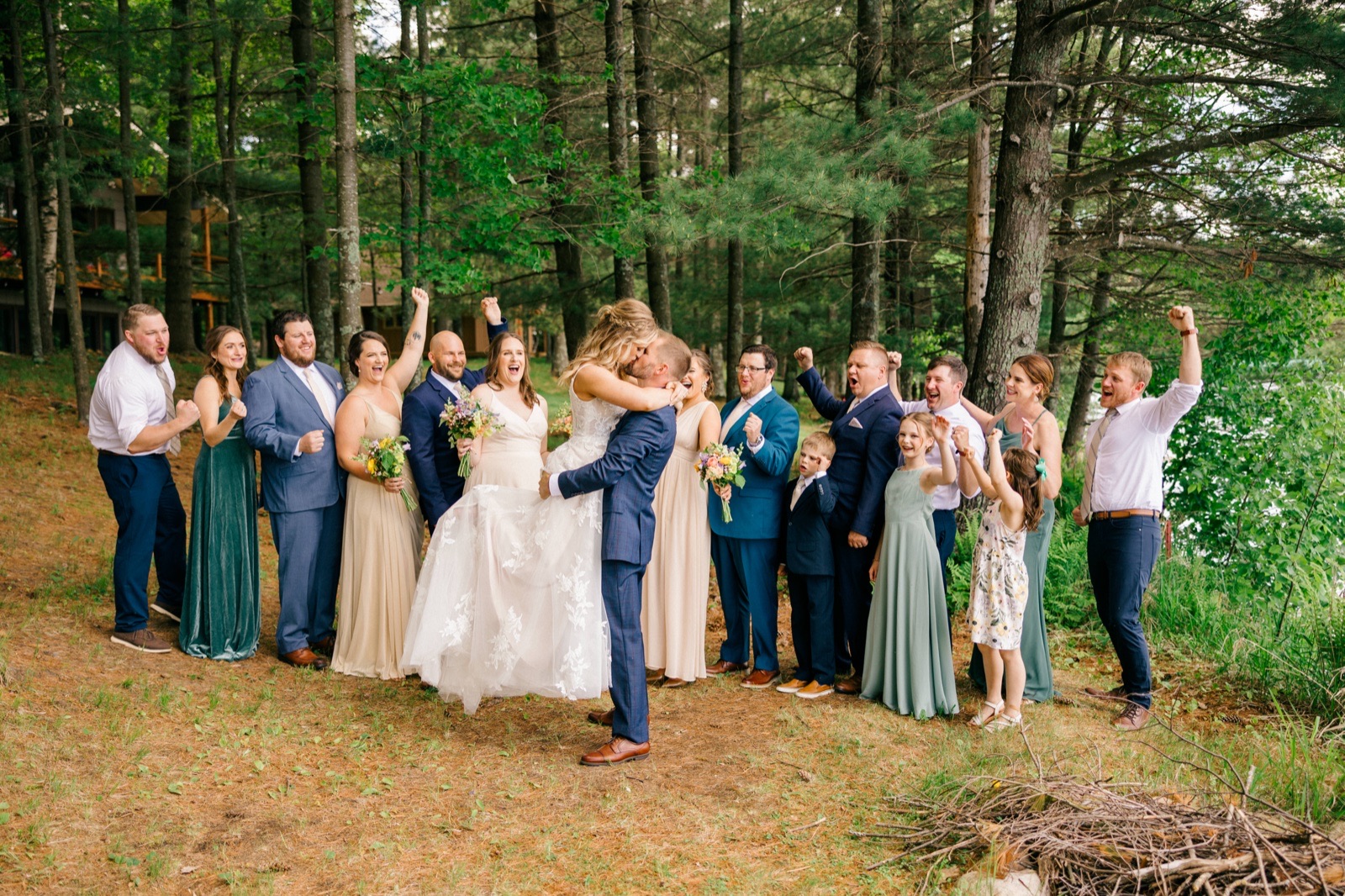 The couple kisses at the center of their bridal party, who cheer with raised fists in a wooded clearing near the lake — Tim Larsen Photography, Brainerd Lakes MN