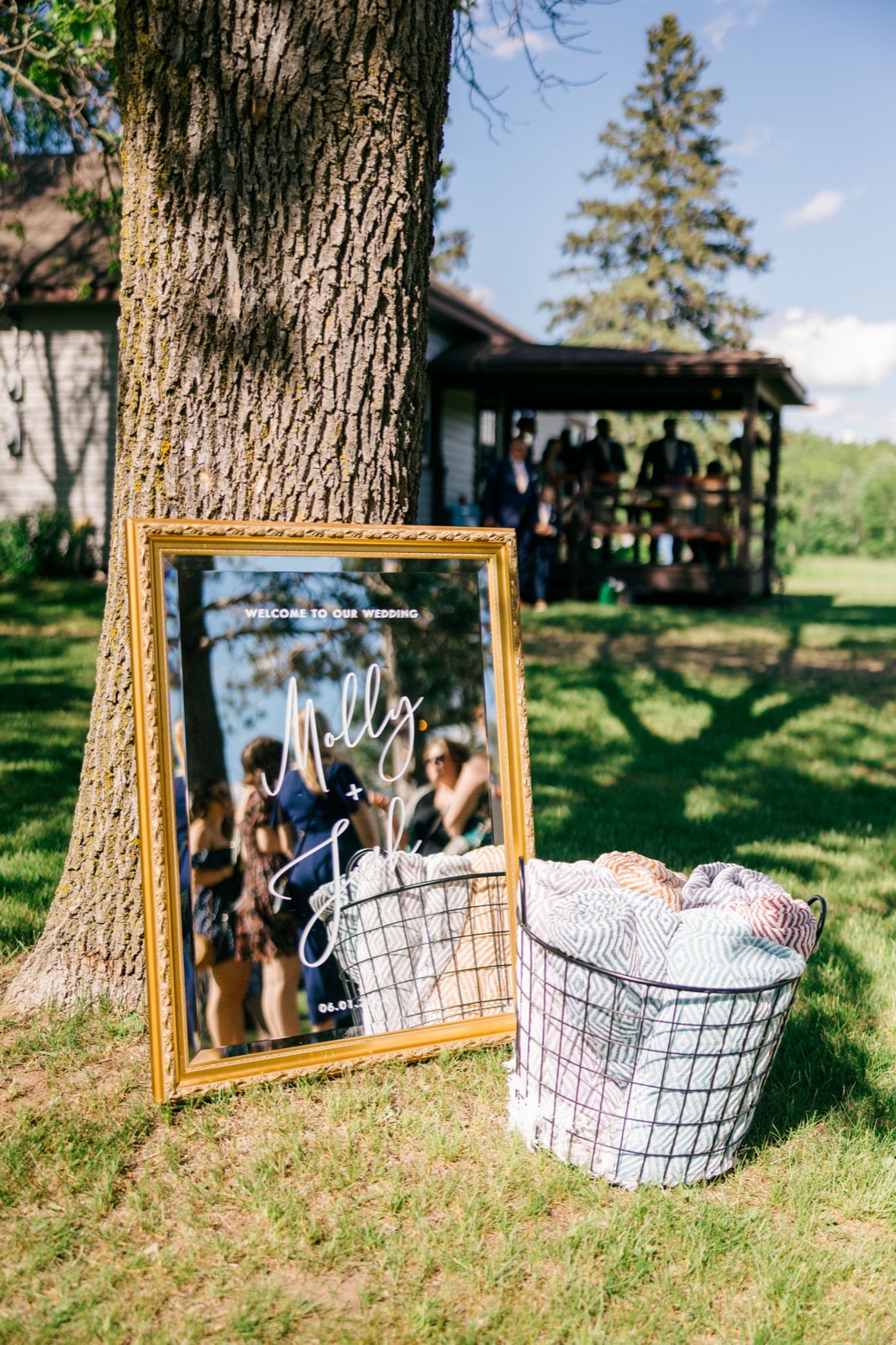 A gold-framed welcome mirror reading 'Molly + Josh' leans against a large oak tree beside a wire basket of folded blankets, with a ceremony pavilion and gathered guests in the background — Tim Larsen Photography, Brainerd Lakes MN