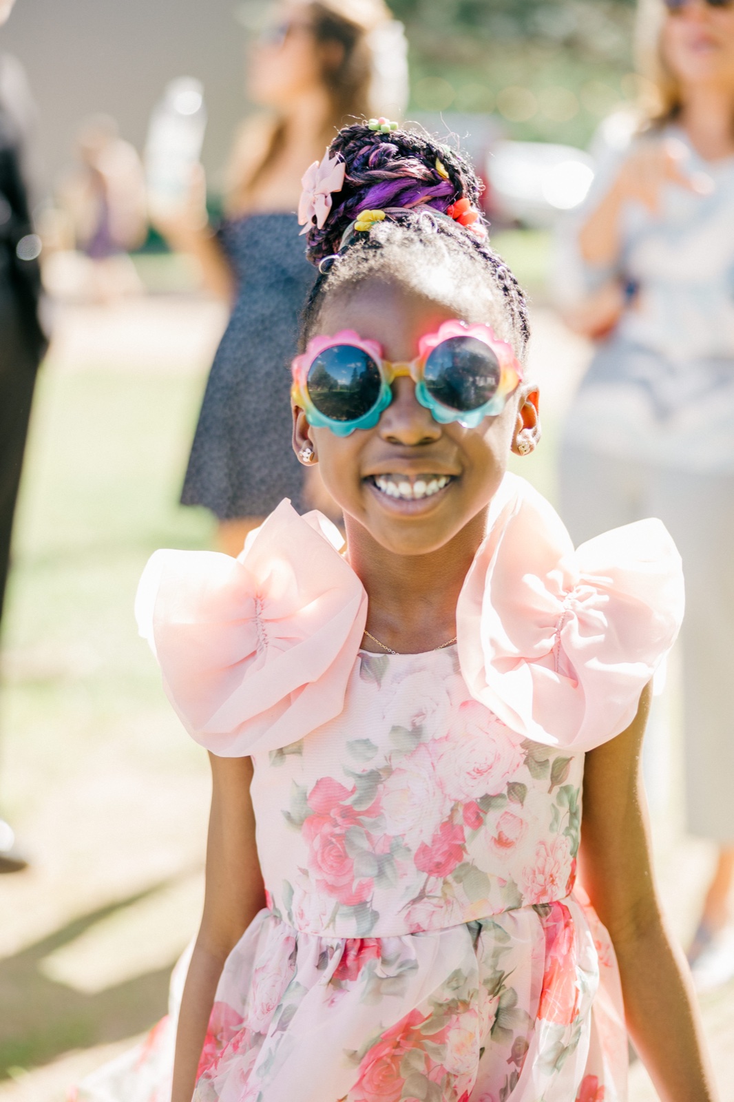 A young flower girl in a pink floral ruffled dress and rainbow flower-shaped sunglasses grins broadly at the camera in bright afternoon sun — Tim Larsen Photography, Brainerd Lakes MN