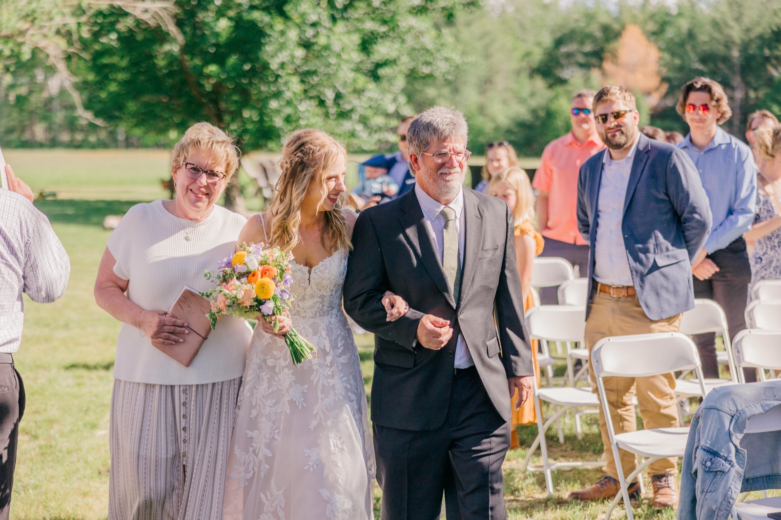 The bride walks down a grass aisle between her parents, smiling and holding a colorful bouquet as seated guests look on — Tim Larsen Photography, Brainerd Lakes MN