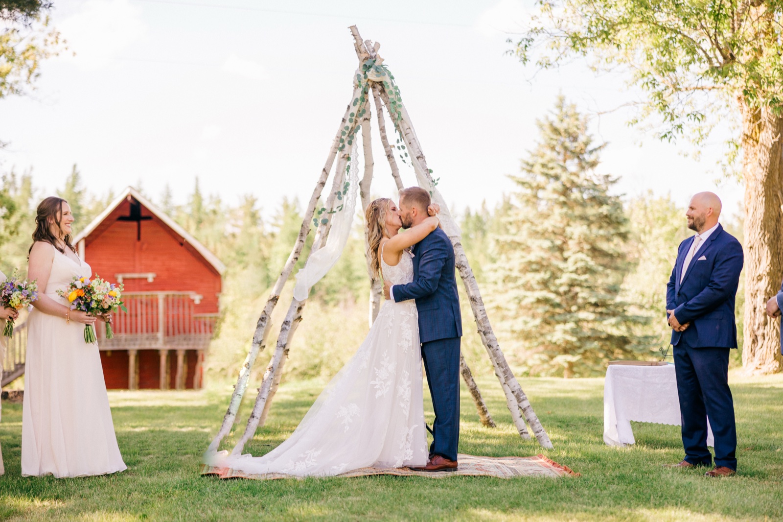 The bride and groom share their first kiss beneath a birch-pole teepee arch on a sunlit lawn, with a bridesmaid holding a wildflower bouquet on the left and a red barn visible in the background — Tim Larsen Photography, Brainerd Lakes MN