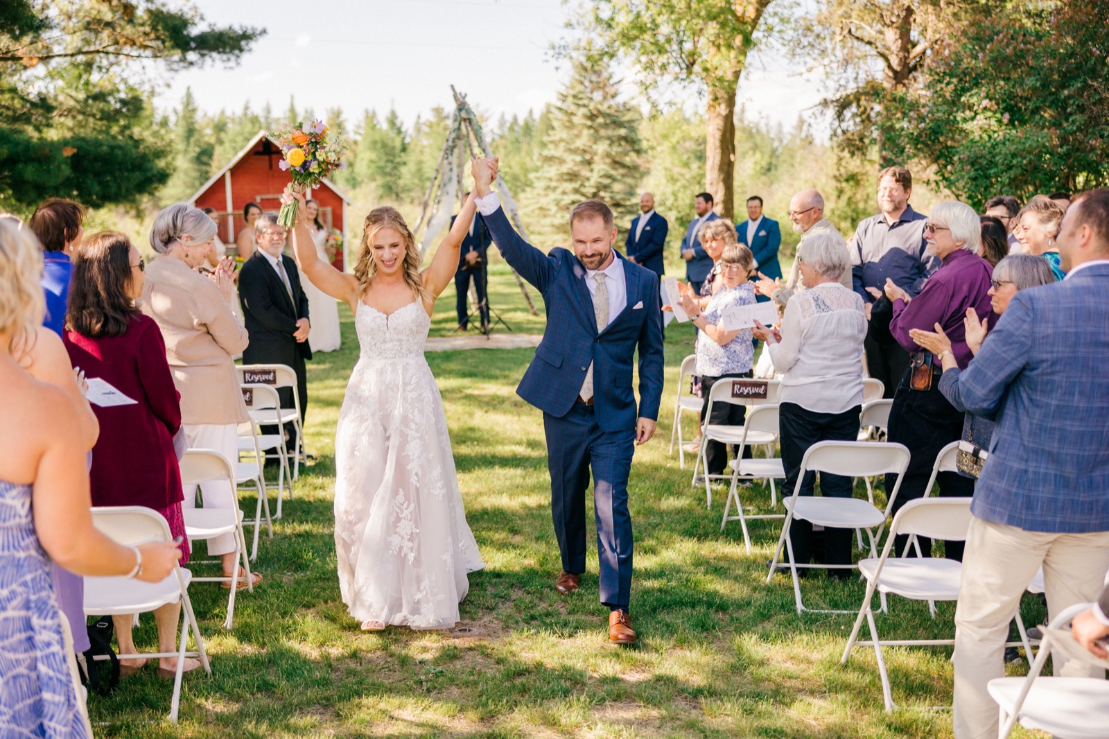 The bride and groom raise their joined hands triumphantly as they walk back down the aisle past clapping guests after their outdoor ceremony — Tim Larsen Photography, Brainerd Lakes MN