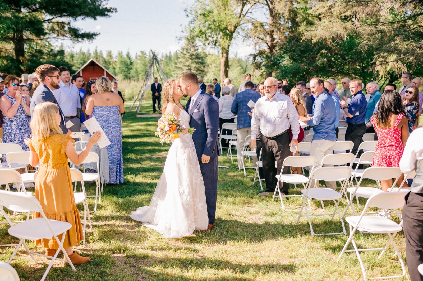 The bride and groom pause to kiss in the middle of the aisle as guests on both sides clap and cheer with a red barn visible in the background — Tim Larsen Photography, Brainerd Lakes MN