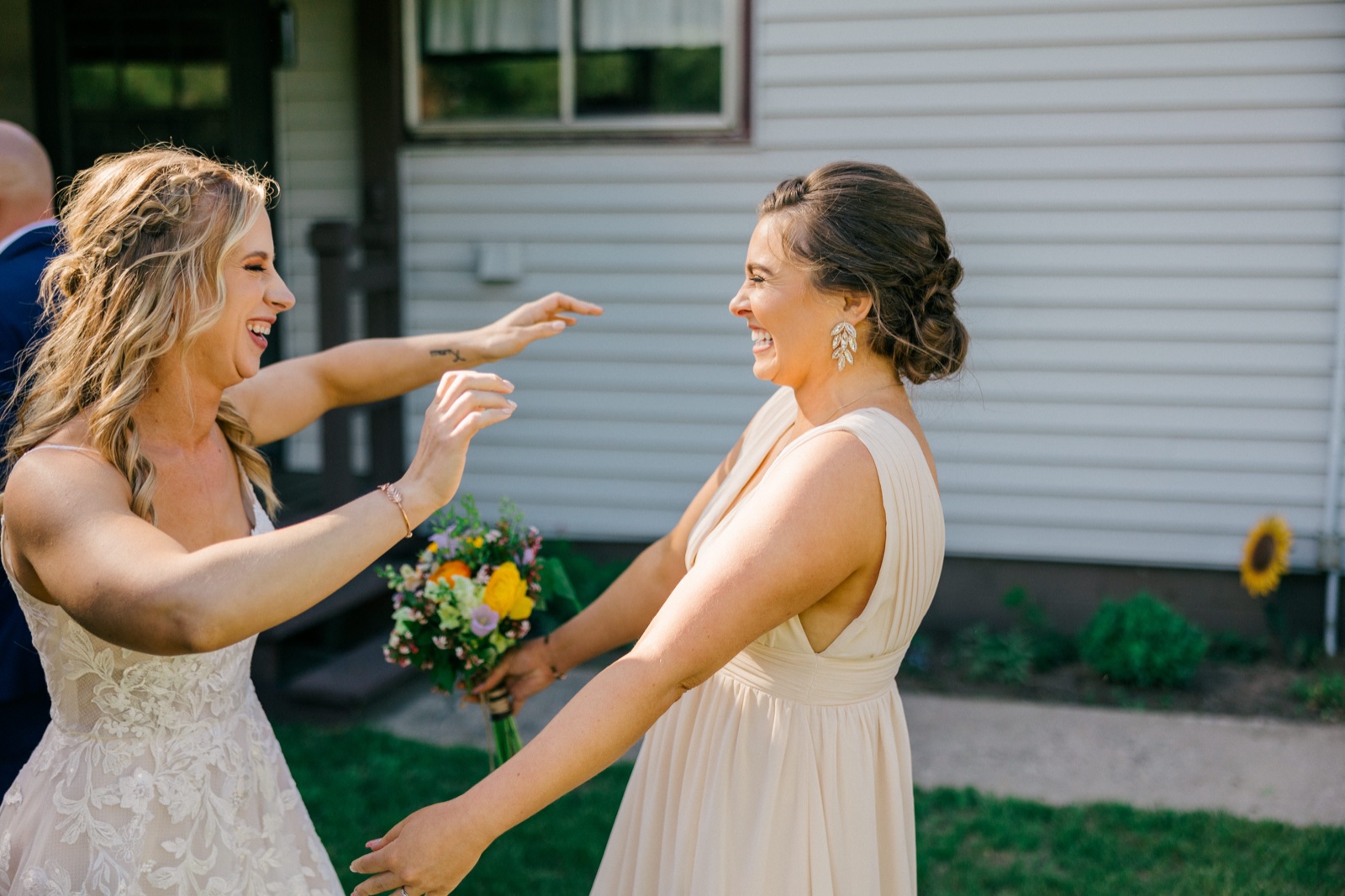 The bride and a bridesmaid laugh and reach toward each other for a hug outside the cabin, the bridesmaid holding a colorful summer bouquet — Tim Larsen Photography, Brainerd Lakes MN