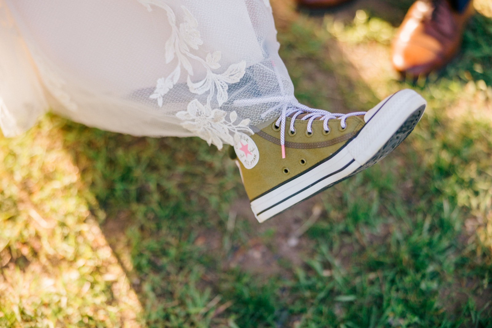 An olive green Converse high-top sneaker peeks out from beneath the bride's lace dress hem as she swings her foot above sunlit grass, the groom's brown dress shoe visible in the background — Tim Larsen Photography, Brainerd Lakes MN