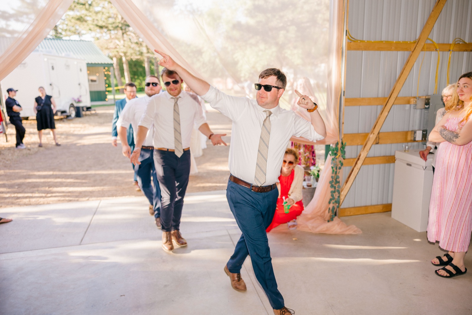 The groom in sunglasses dances through a backlit drapery entrance with arms raised, leading the groomsmen into the reception while a seated guest watches and laughs — Tim Larsen Photography, Brainerd Lakes MN