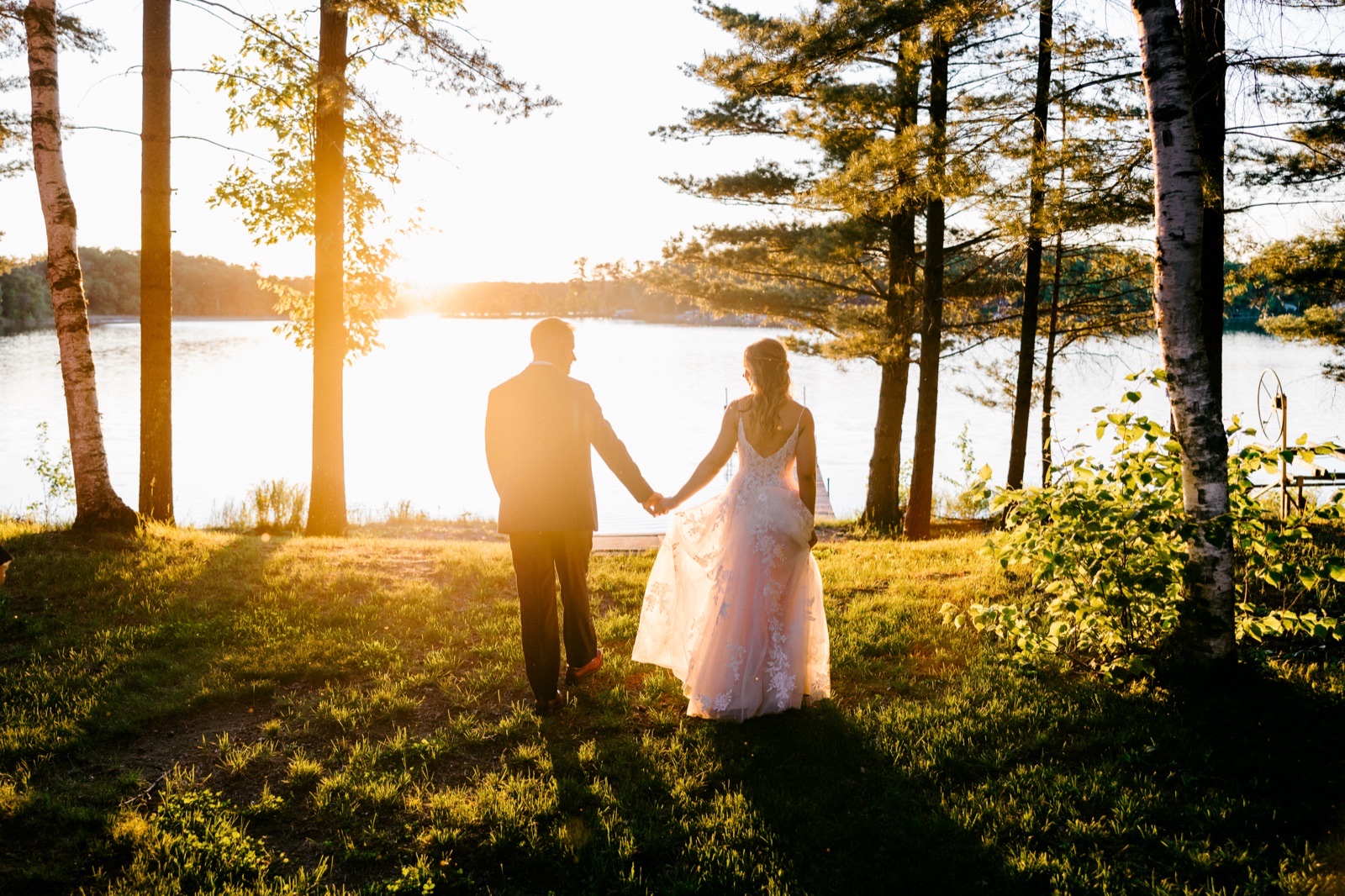 The bride and groom walk hand in hand toward the lake at sunset, framed by tall birch and pine trees with the sun flaring behind them — Tim Larsen Photography, Brainerd Lakes MN