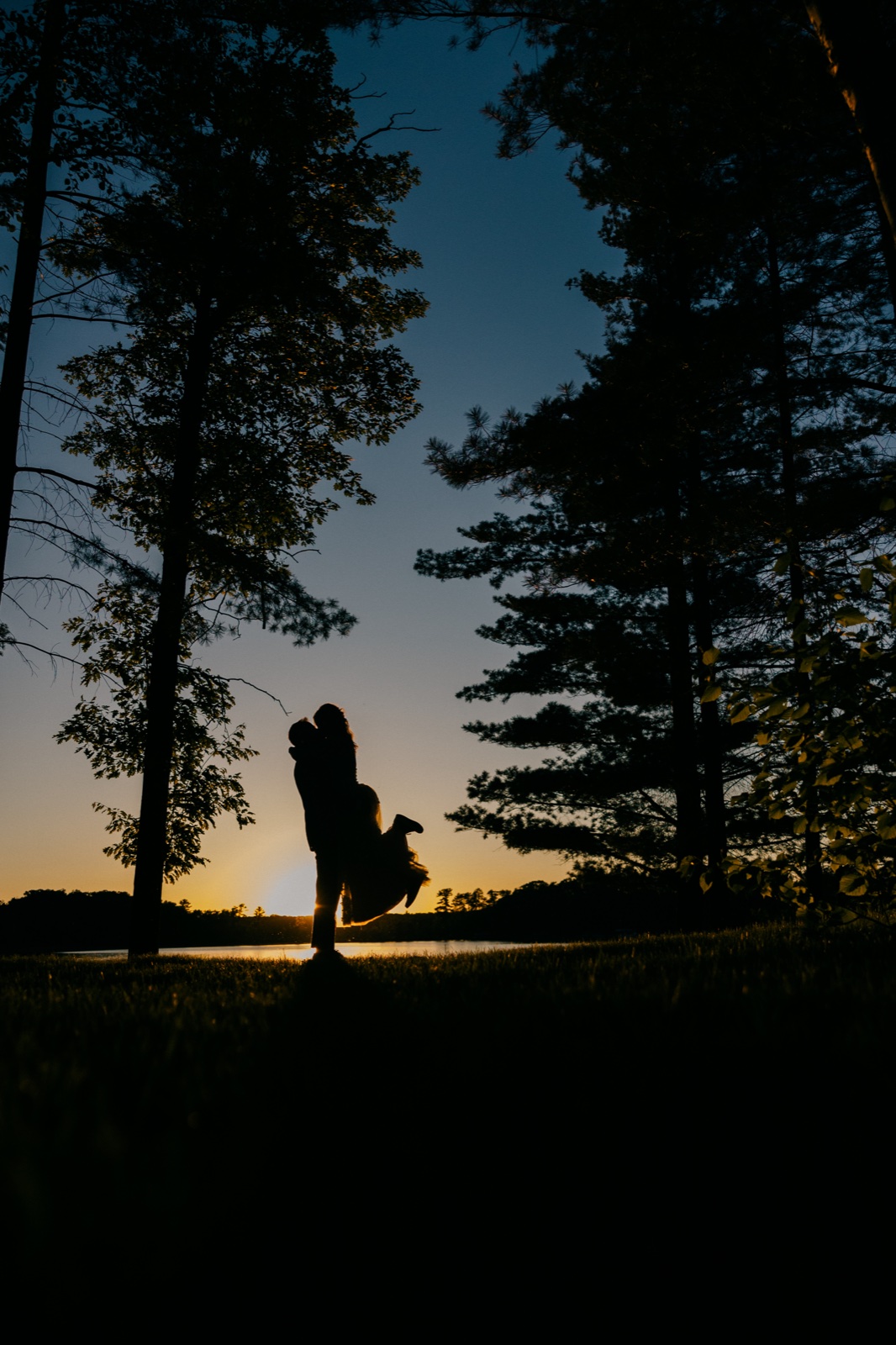 The groom lifts the bride in a sunset silhouette on the lakeshore lawn, framed by tall pine trees with the sun setting over the water behind them — Tim Larsen Photography, Brainerd Lakes MN