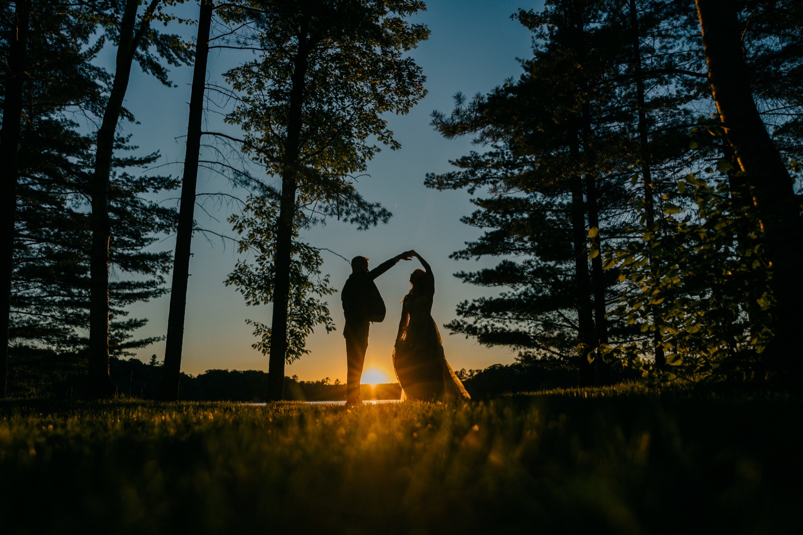 The couple dance in silhouette against a glowing sunset between tall pines at the edge of the lake — Tim Larsen Photography, Brainerd Lakes MN