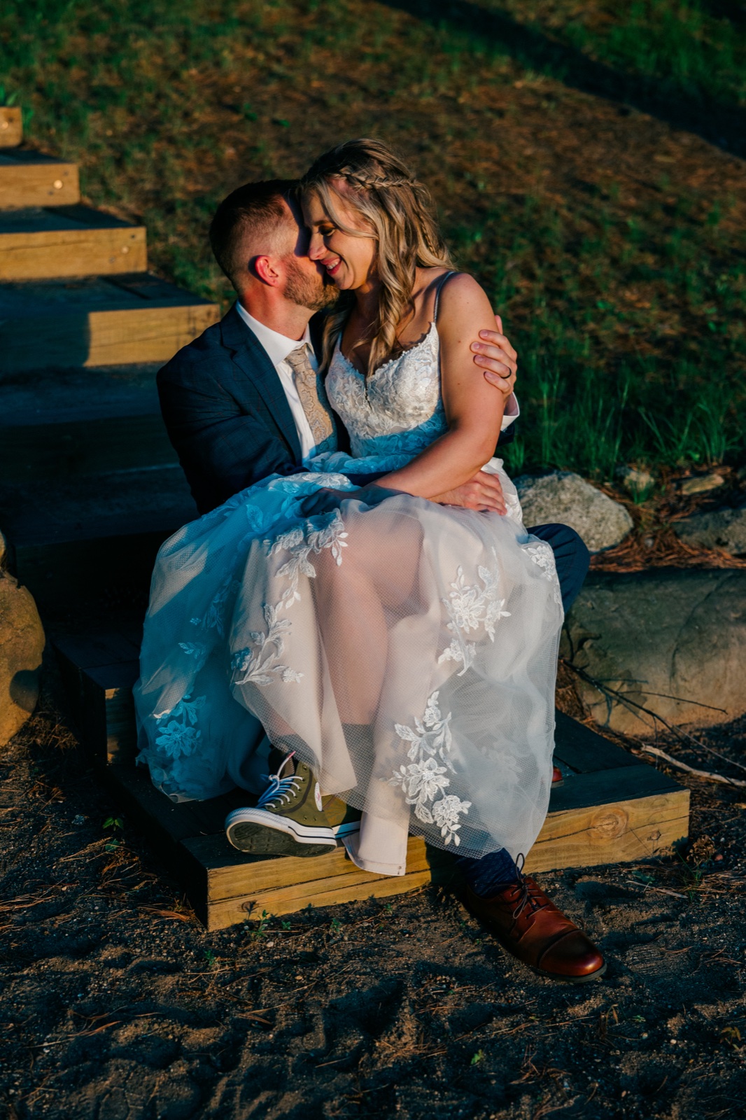 The bride and groom share a quiet kiss while seated on wooden steps at golden hour, her lace dress spilling over the edge and his green sneakers visible beneath — Tim Larsen Photography, Brainerd Lakes MN
