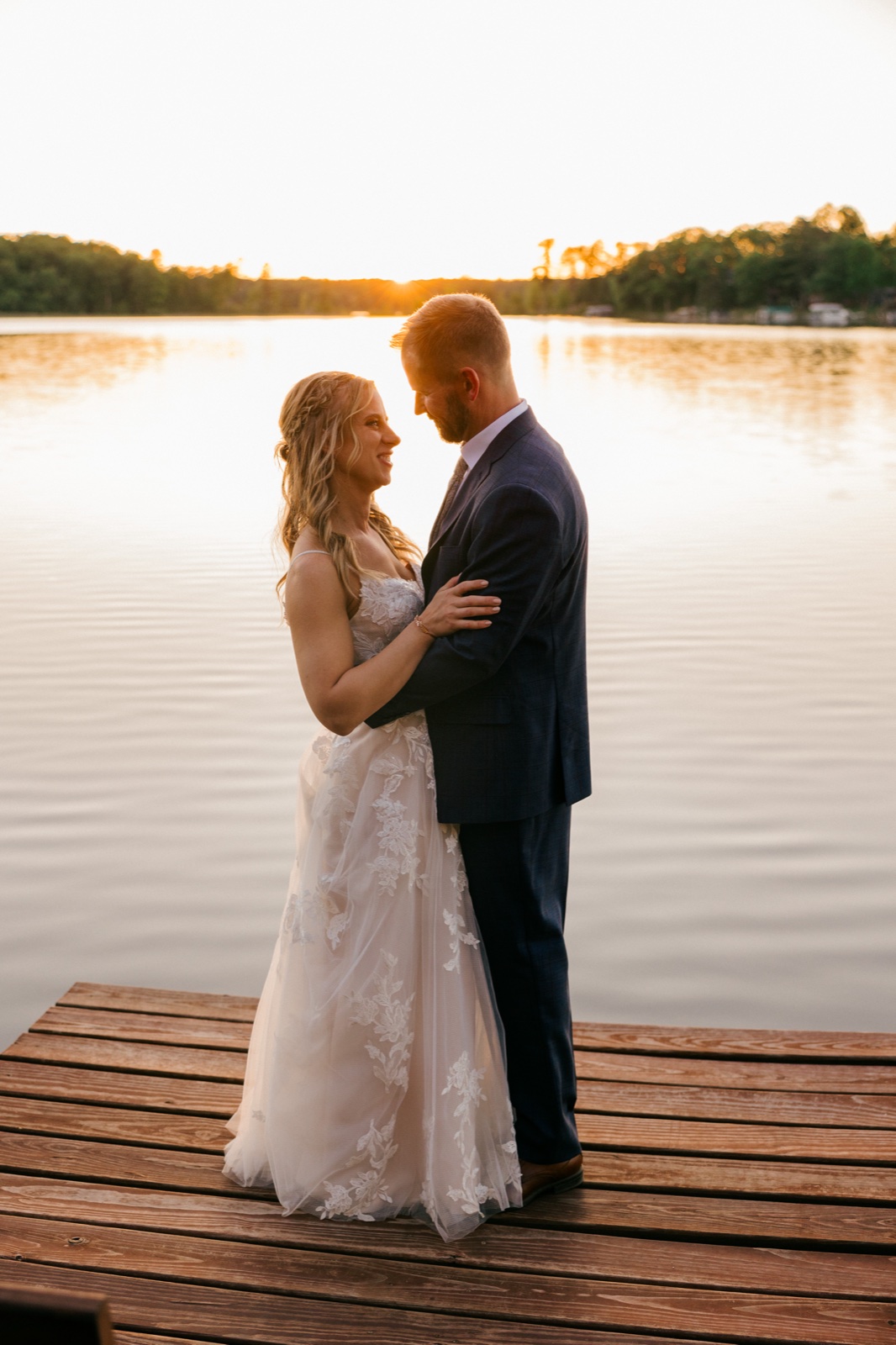 The bride and groom stand forehead-to-forehead on the wooden dock as the sun sets behind them over the calm Minnesota lake — Tim Larsen Photography, Brainerd Lakes MN