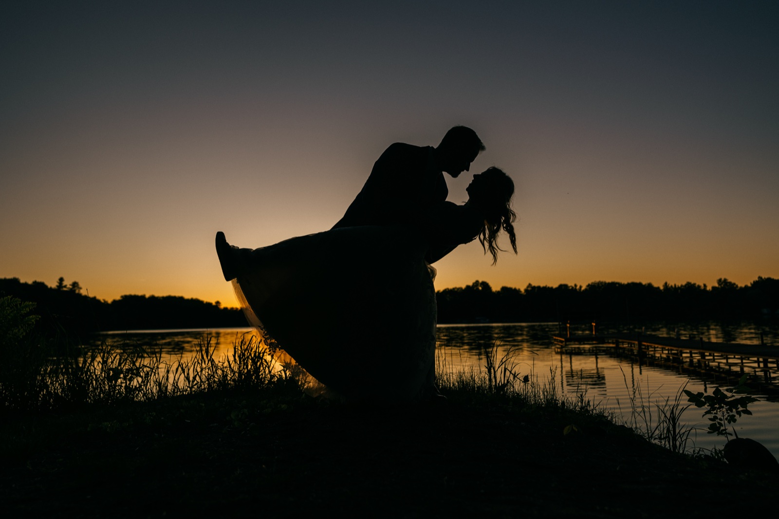 The groom dips the bride into a near-kiss as their silhouettes stand against a glowing dusk sky over a Brainerd Lakes lakeshore with a dock extending into the water — Tim Larsen Photography, Brainerd Lakes MN