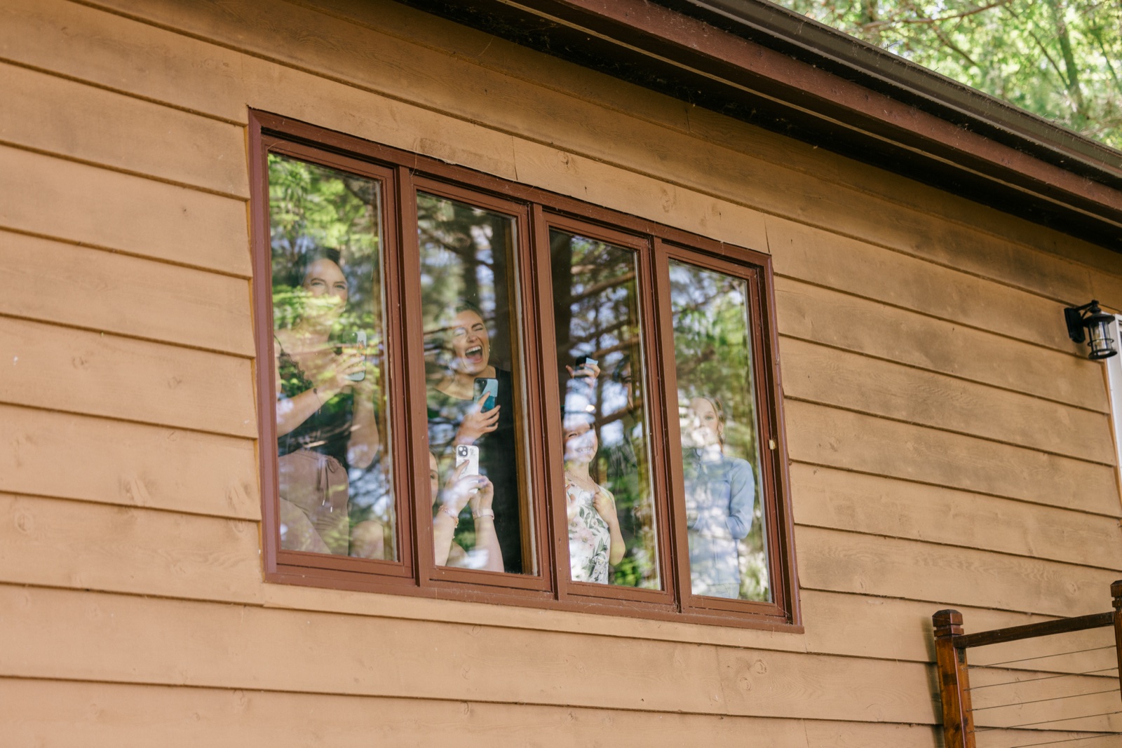 Viewed from outside the cabin, the bride and her bridesmaids laugh and hold up phones in a row of windows along the wood-sided wall — Tim Larsen Photography, Brainerd Lakes MN