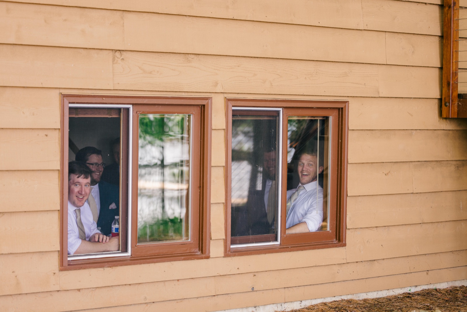 Four groomsmen in shirts and ties peer out through two cabin windows, smiling and laughing at the camera — Tim Larsen Photography, Brainerd Lakes MN
