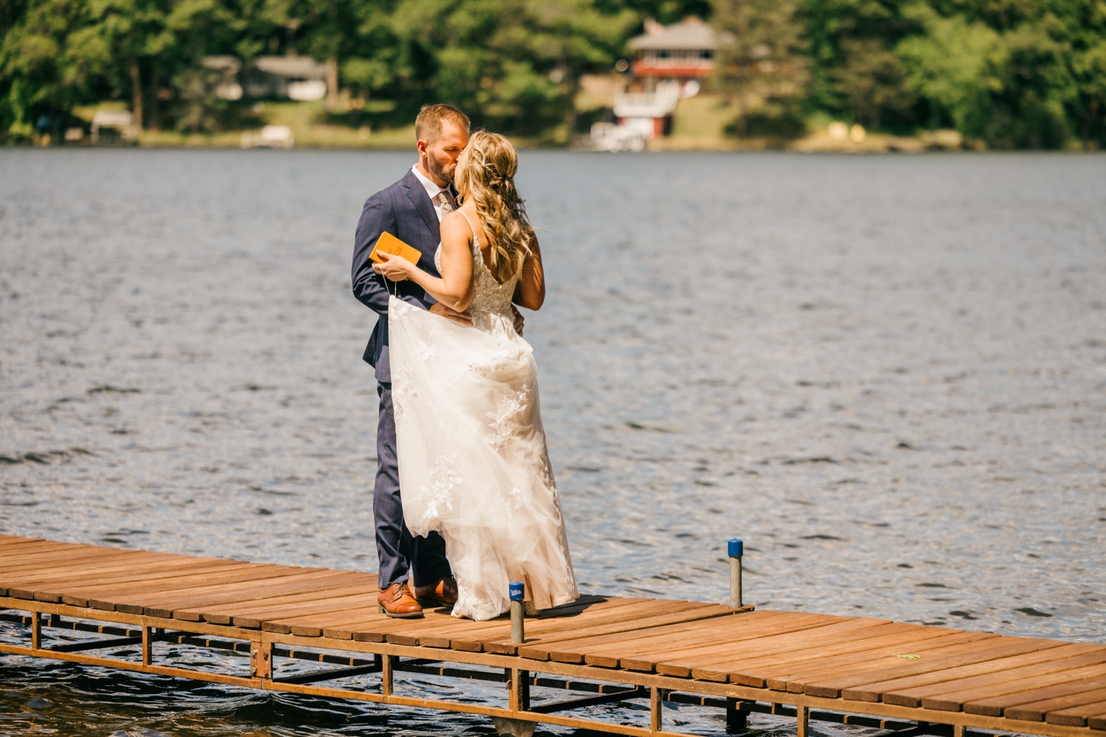 The bride and groom kiss at the end of the wooden dock extending into the lake, the groom holding a small vow book during their private vows — Tim Larsen Photography, Brainerd Lakes MN