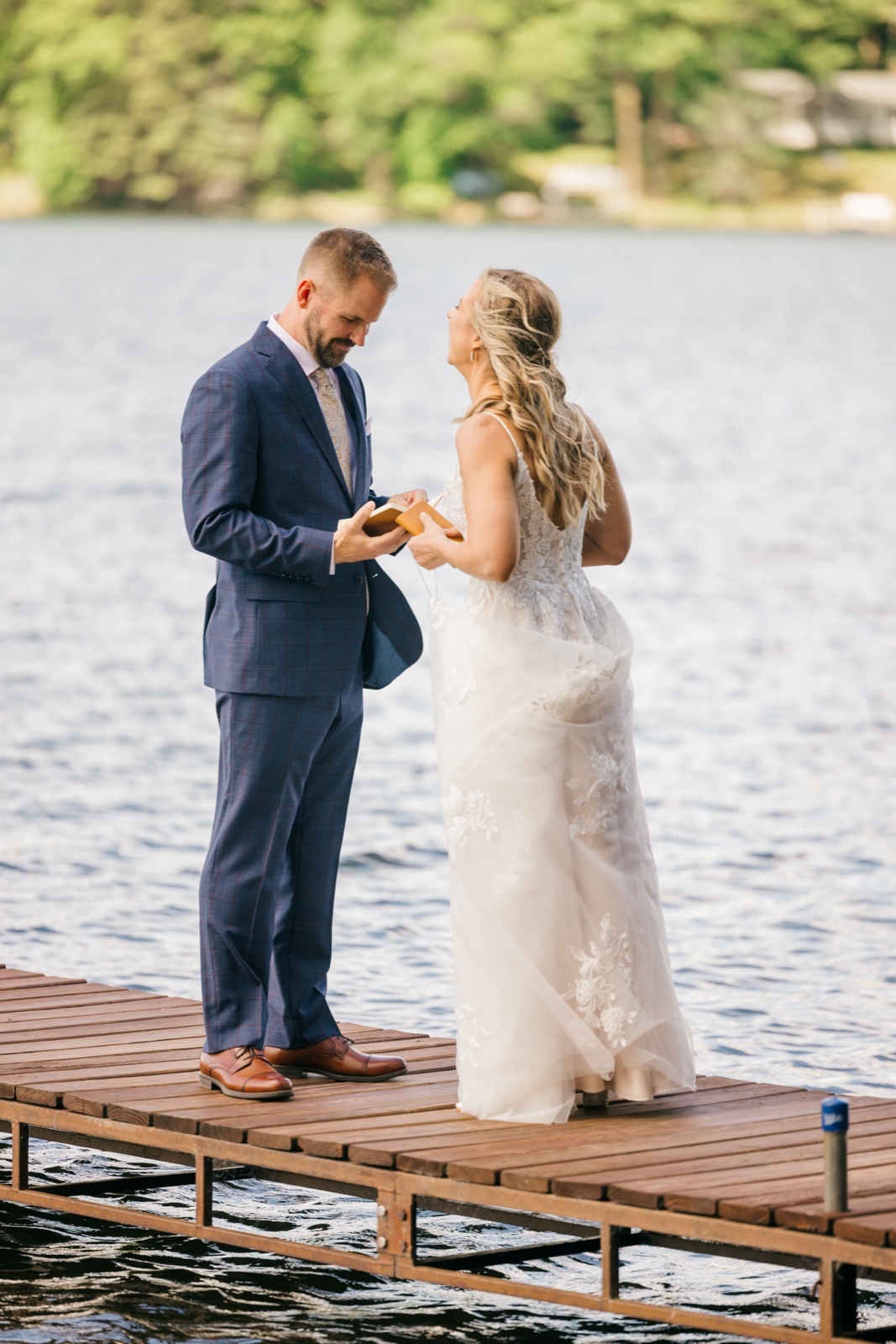 The groom reads from a small vow book while the bride stands facing him on the wooden dock over the lake during their private vows — Tim Larsen Photography, Brainerd Lakes MN