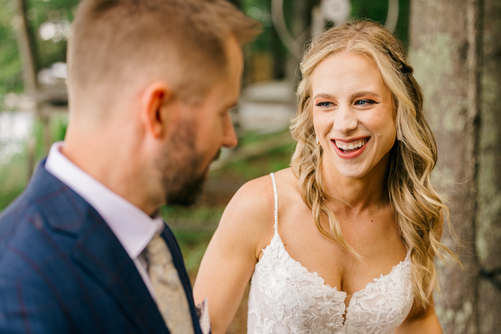 The bride laughs openly at the groom during a first look in a wooded clearing, his out-of-focus profile framing the left side of the image — Tim Larsen Photography, Brainerd Lakes MN