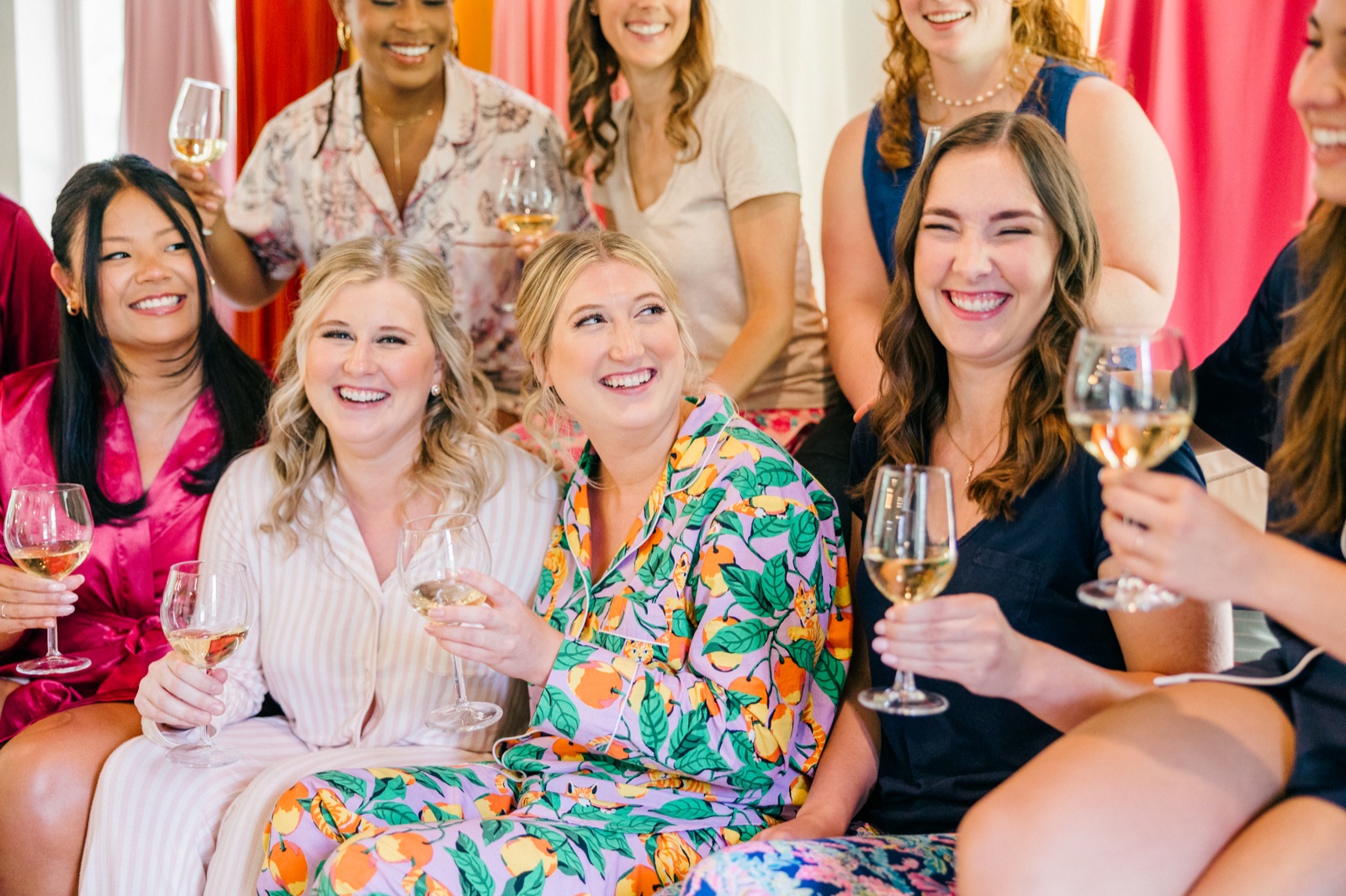 Monika sits laughing in matching pajamas surrounded by her bridesmaids holding raised wine glasses against a pink fabric backdrop — Tim Larsen Photography, Brainerd Lakes MN