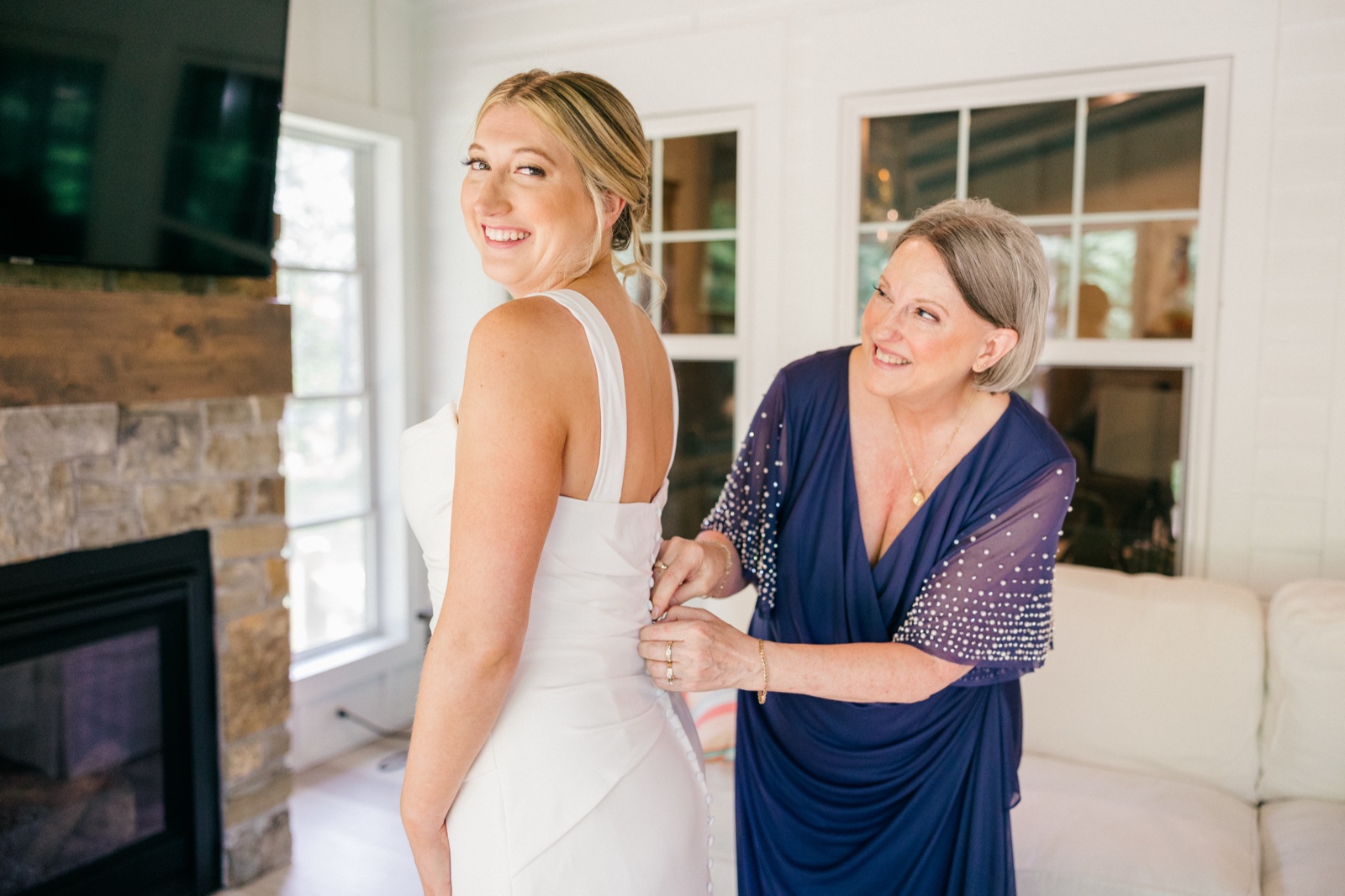 Monika glances over her shoulder with a wide smile as her mother fastens the buttons on the back of her white gown — Tim Larsen Photography, Brainerd Lakes MN