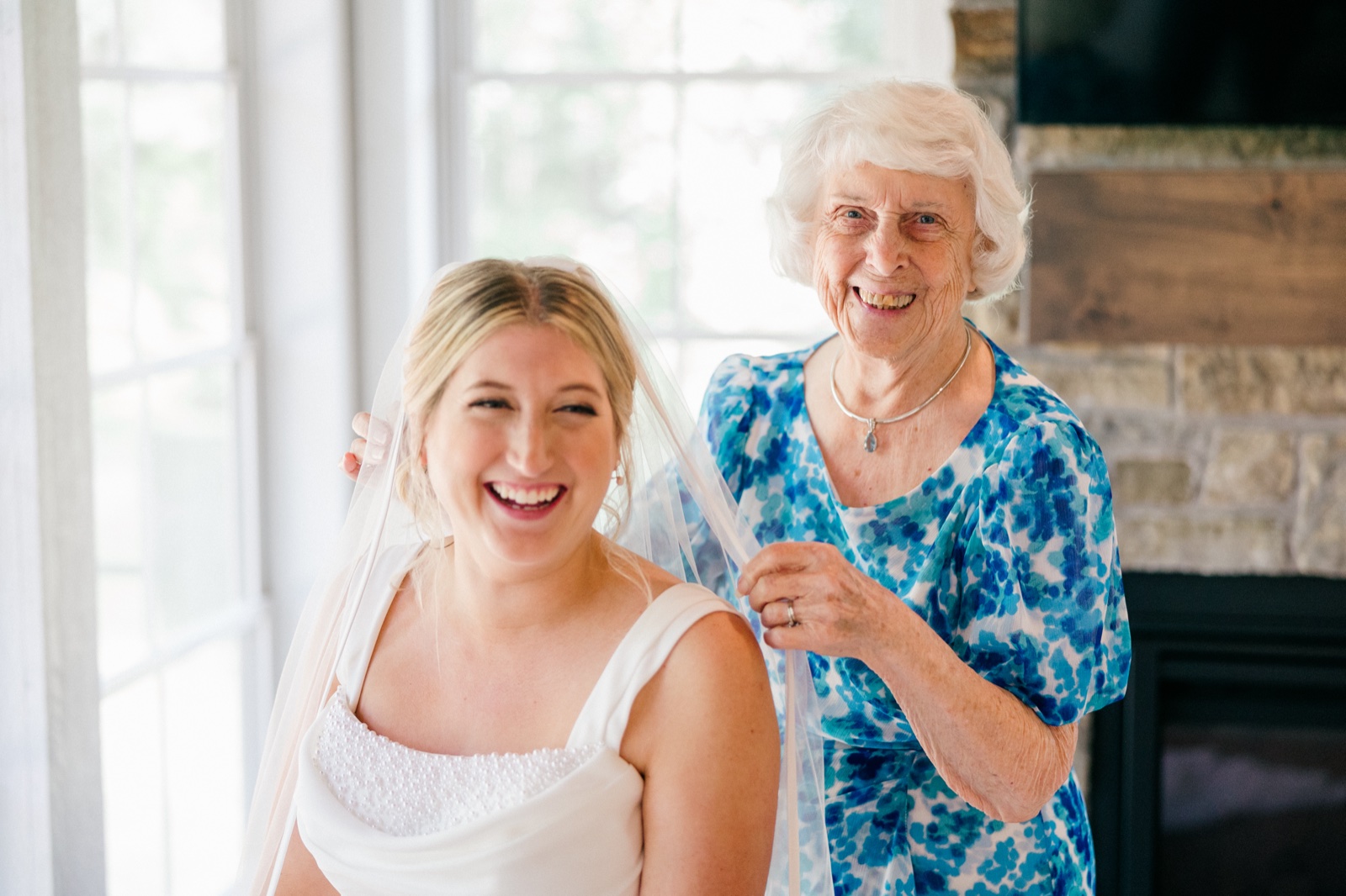 Monika laughs in her wedding dress and veil while her grandmother smiles broadly behind her in a window-lit room — Tim Larsen Photography, Brainerd Lakes MN
