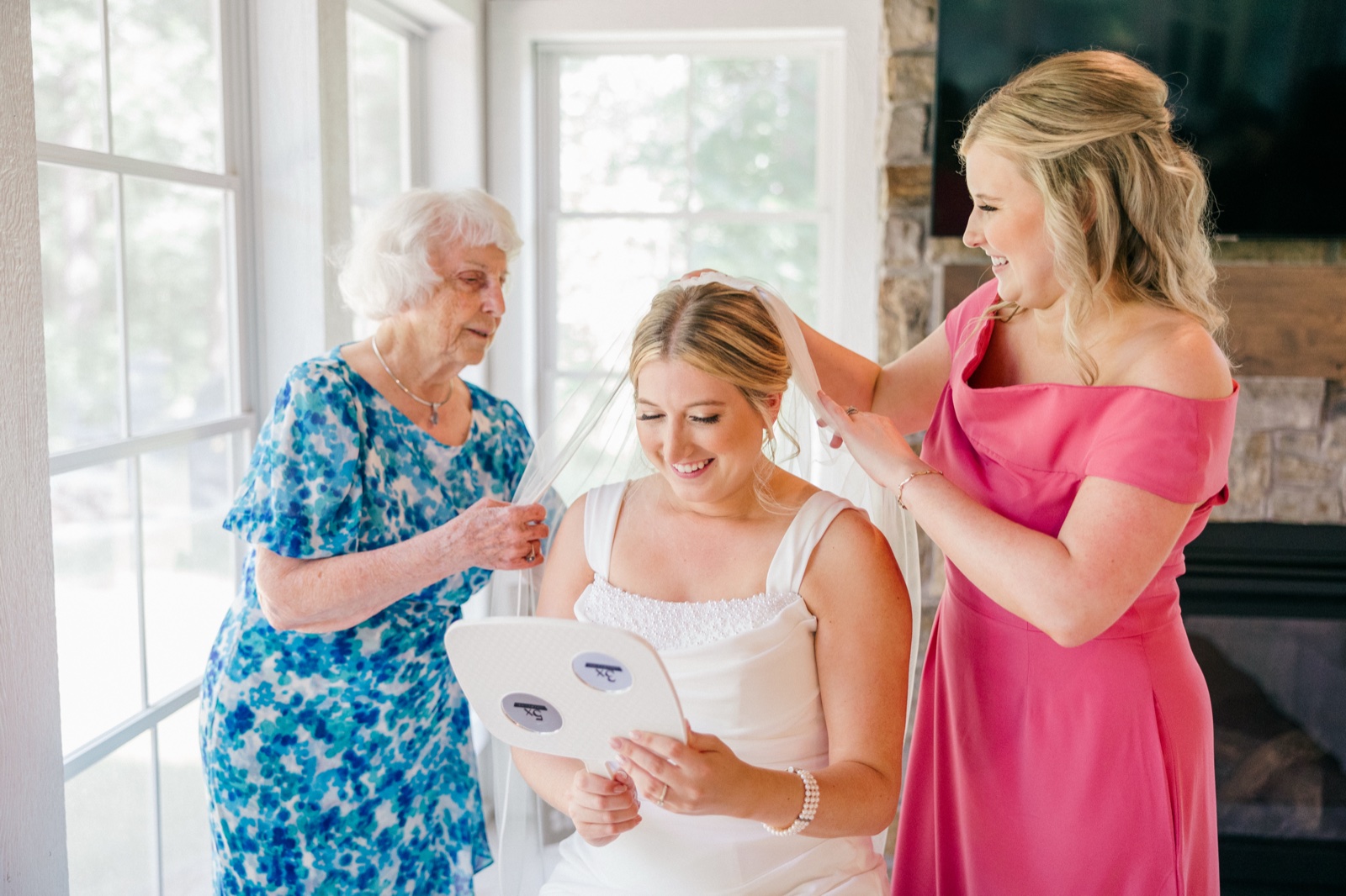 The bride looks down at a hand mirror smiling while her grandmother and a bridesmaid help adjust her veil — Tim Larsen Photography, Brainerd Lakes MN