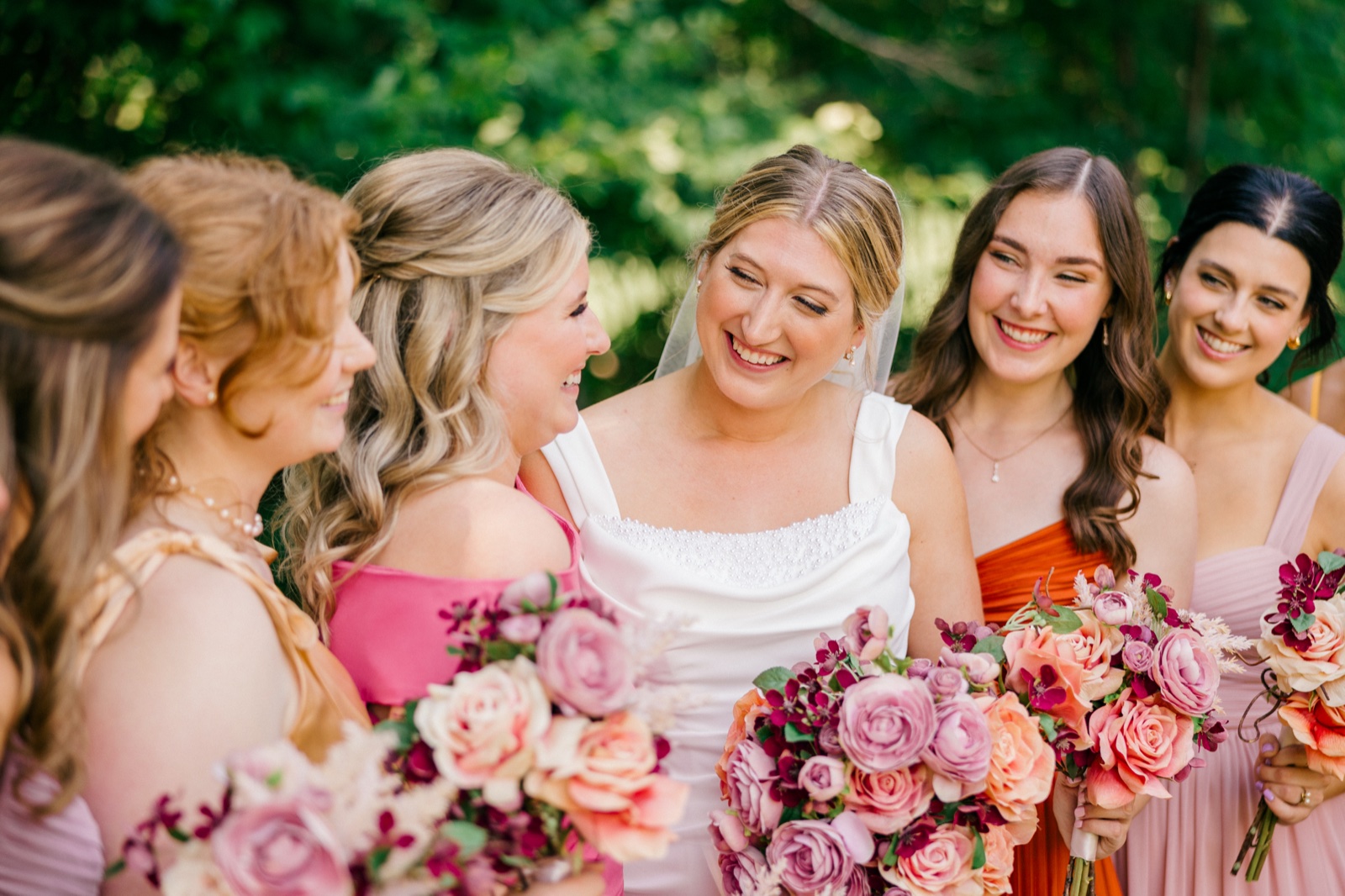 Monika laughs with her bridesmaids gathered close around her, holding pink and coral bouquets in front of summer greenery — Tim Larsen Photography, Brainerd Lakes MN