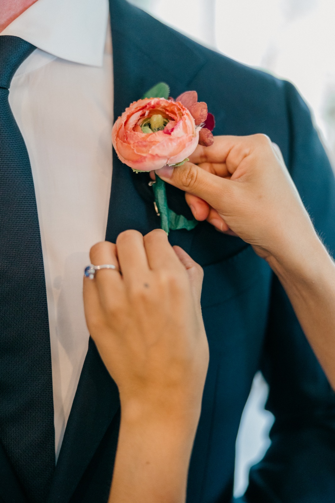 Close-up of the bride's hands pinning a coral ranunculus boutonniere onto the groom's navy lapel — Tim Larsen Photography, Brainerd Lakes MN