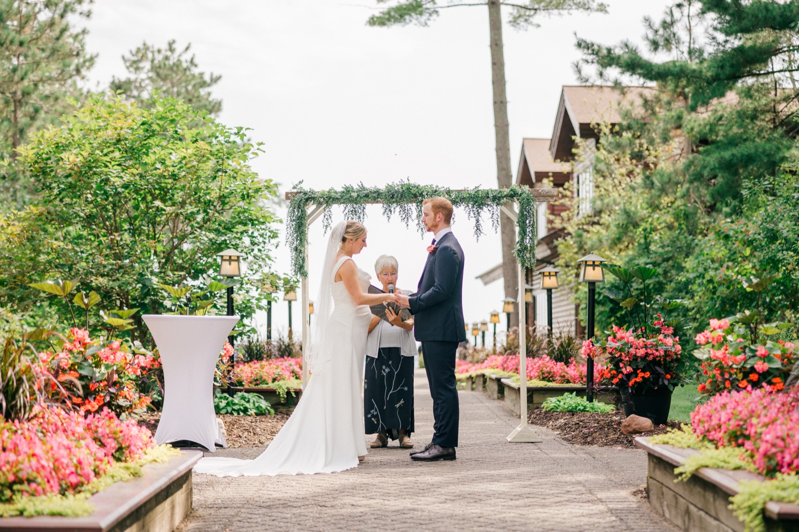 Monika and Mats hold hands beneath a greenery-draped arch as the officiant reads, lush flowerbeds and lanterns lining the outdoor aisle at Grand View Lodge — Tim Larsen Photography, Brainerd Lakes MN