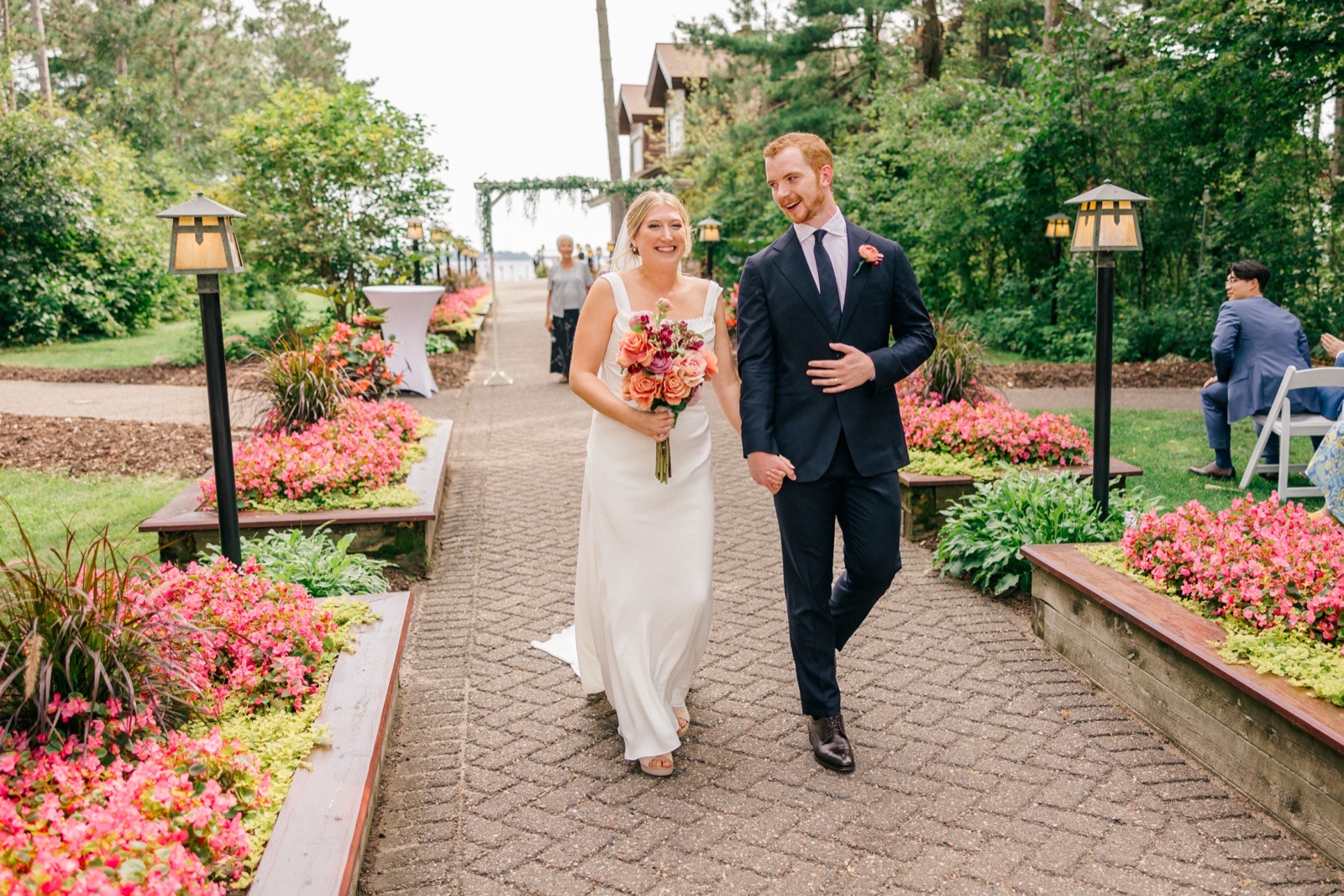 Monika and Mats laugh as they walk down a brick path lined with pink flowerbeds and lanterns just after their lakeside ceremony at Grand View Lodge — Tim Larsen Photography, Brainerd Lakes MN