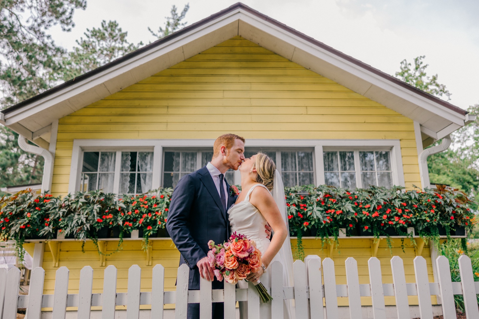 Monika and Mats kiss behind a white picket fence in front of a yellow cottage at Grand View Lodge, the bride holding a coral and pink bouquet — Tim Larsen Photography, Brainerd Lakes MN