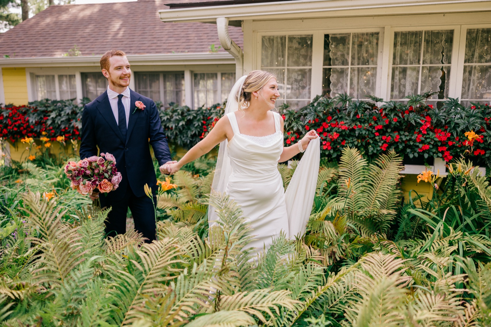 The bride lifts her veil as she walks hand-in-hand with the groom through tall ferns past a Grand View Lodge cottage — Tim Larsen Photography, Brainerd Lakes MN