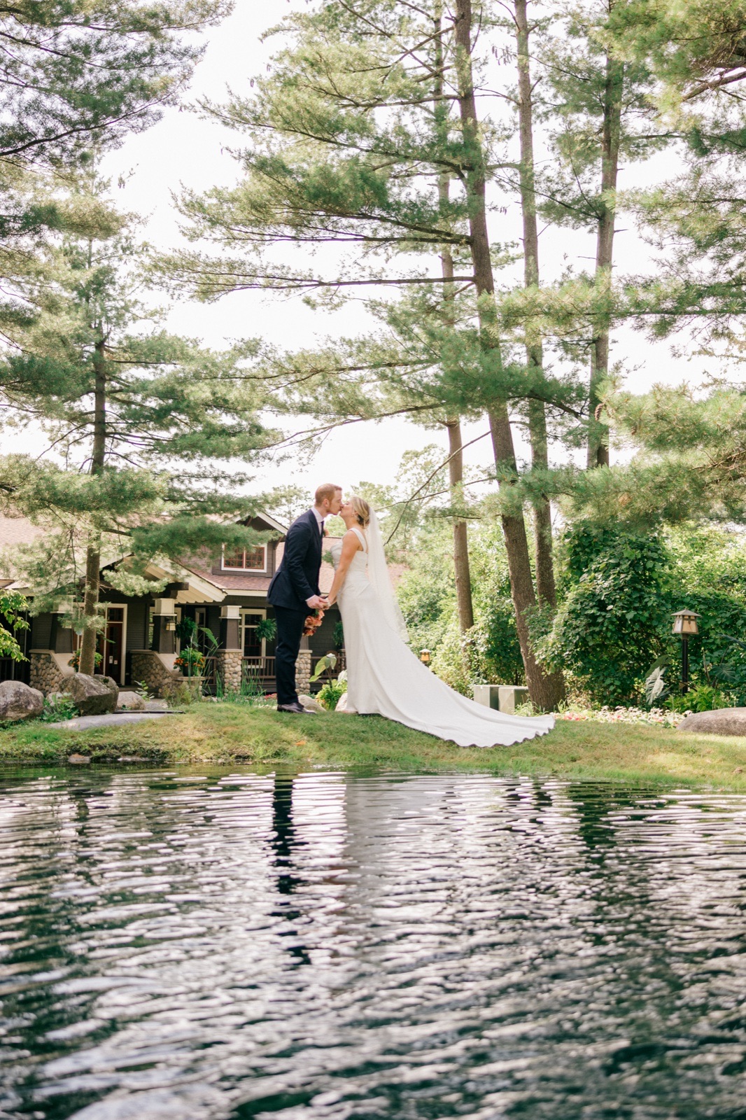 Monika and Mats stand together on a lawn beside a pond, the bride's long train trailing across the grass beneath towering pine trees — Tim Larsen Photography, Brainerd Lakes MN