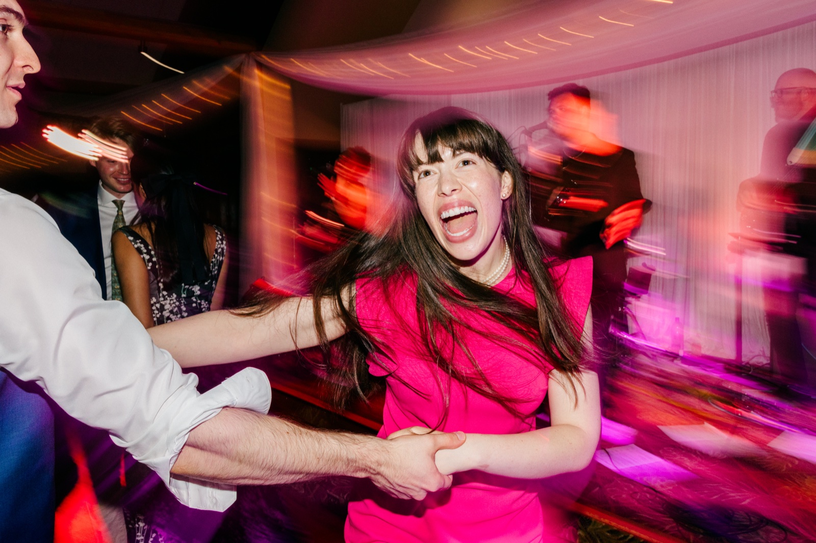 A guest in a hot pink dress laughs open-mouthed mid-spin on the dance floor while holding hands with a partner, surrounded by motion-blurred magenta light streaks — Tim Larsen Photography, Brainerd Lakes MN