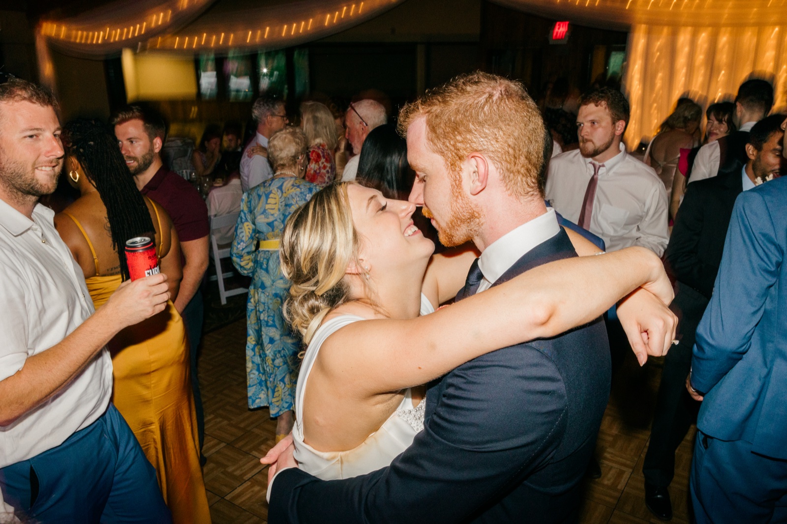 Monika and Mats press their faces close together, laughing, while guests dance and mingle around them under warm string lights — Tim Larsen Photography, Brainerd Lakes MN