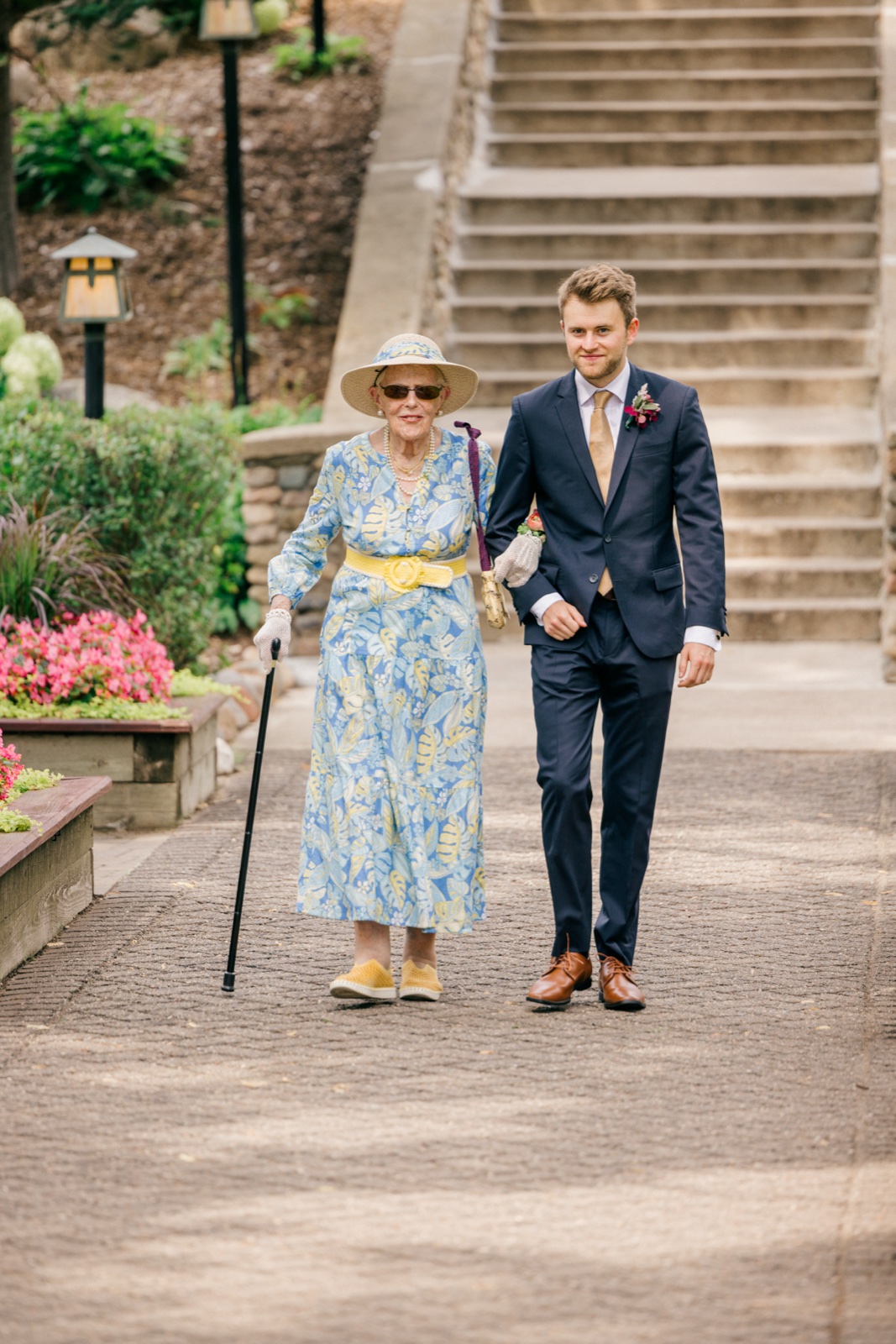 The groom escorts an older woman in a floral dress and sun hat down a gravel path past flower beds at Grand View Lodge — Tim Larsen Photography, Brainerd Lakes MN