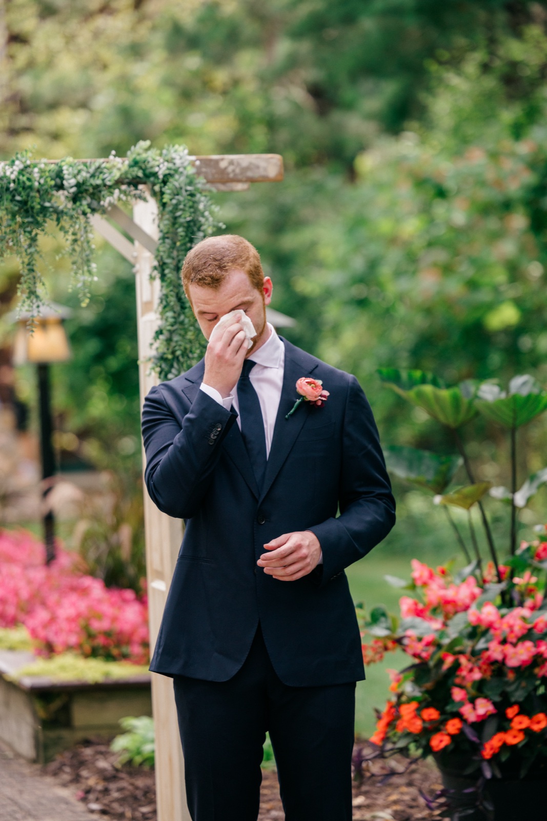 Mats raises a handkerchief to his eyes beneath a greenery-covered ceremony arch surrounded by pink and coral flowers — Tim Larsen Photography, Brainerd Lakes MN