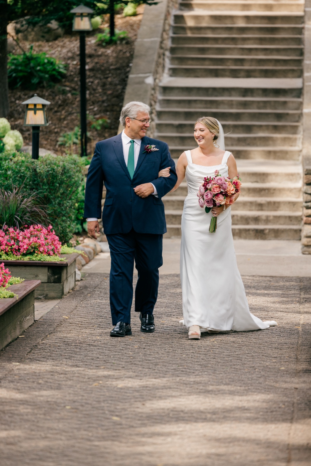 Monika laughs with her father as they walk arm-in-arm down a path with Grand View Lodge's stone staircase rising behind them — Tim Larsen Photography, Brainerd Lakes MN